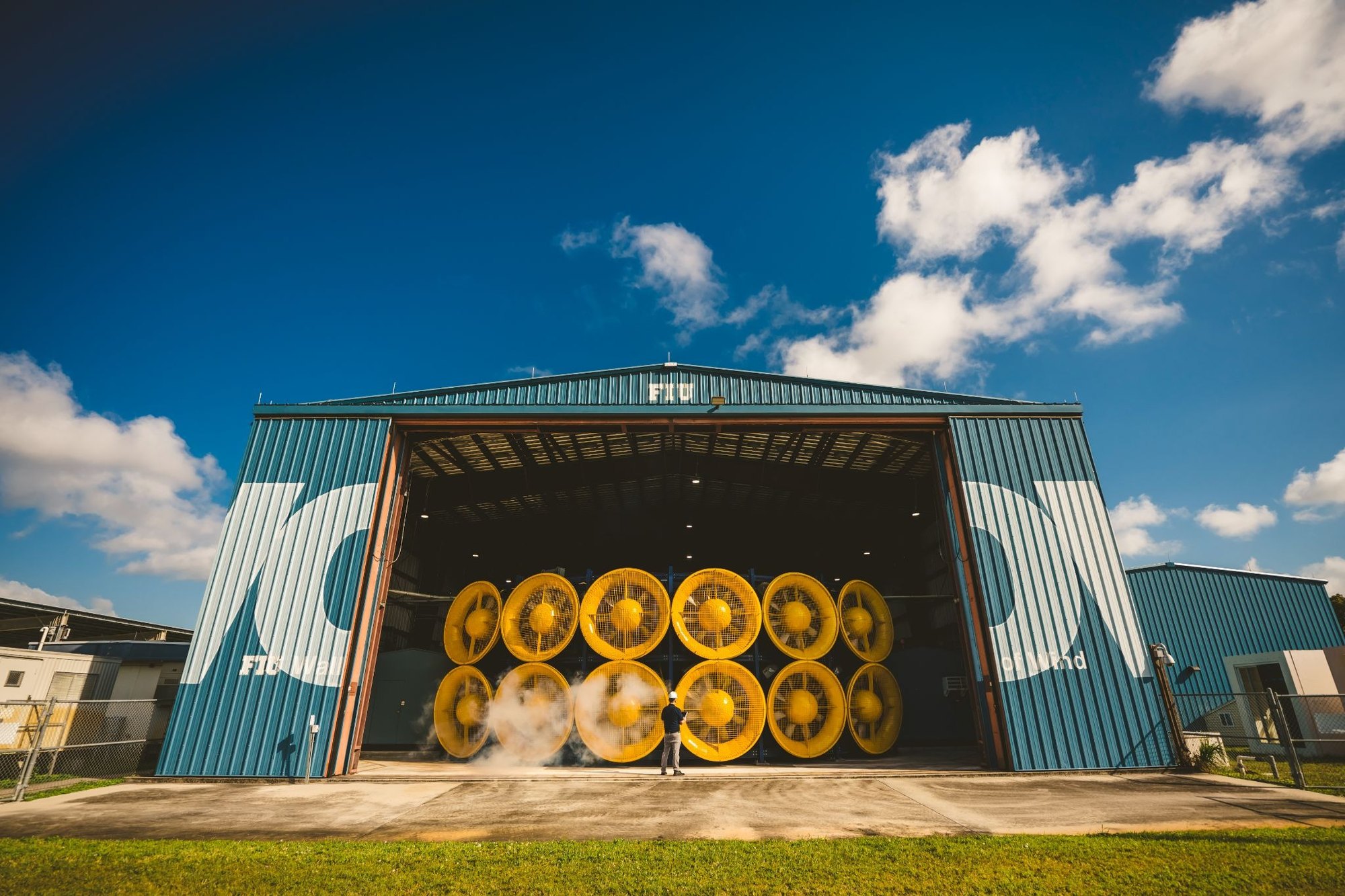 Twelve large yellow industrial fans arranged in a grid inside a blue FIU building, with a person standing in front for scale.