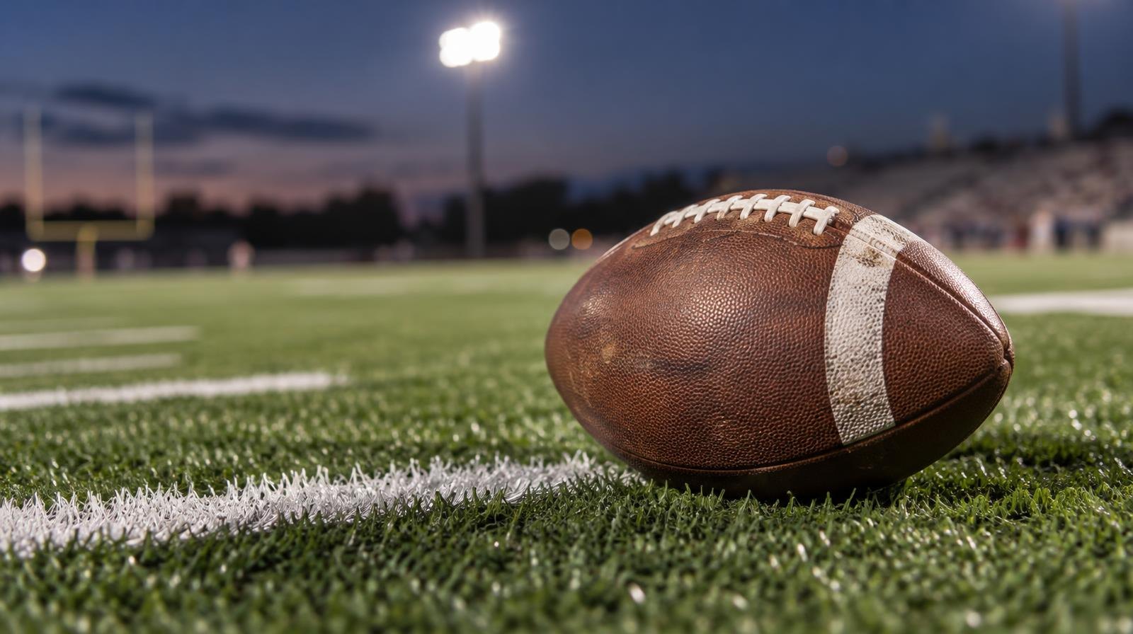 An American football resting on green turf beside a painted white yard line.