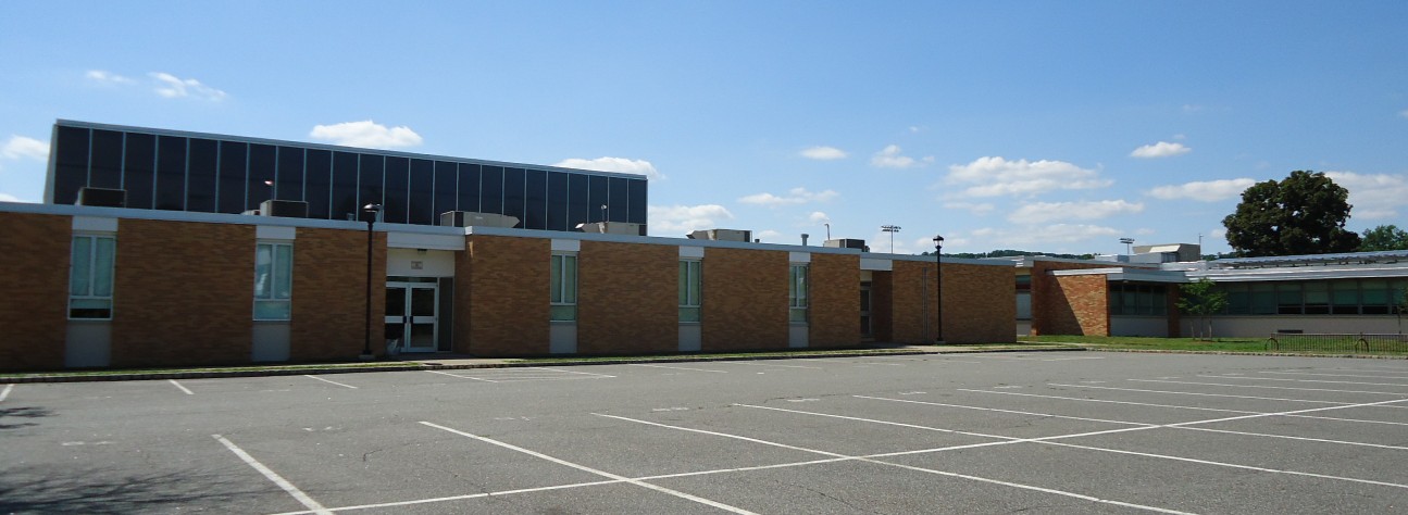 Brick school building exterior with a large parking lot under a blue sky.