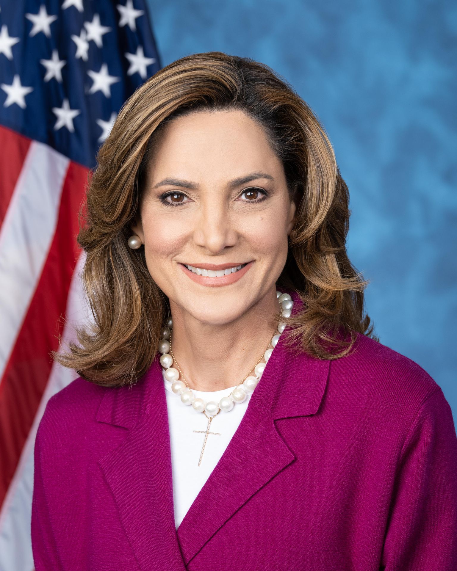 Woman in magenta blazer with pearl necklace posed before an American flag.