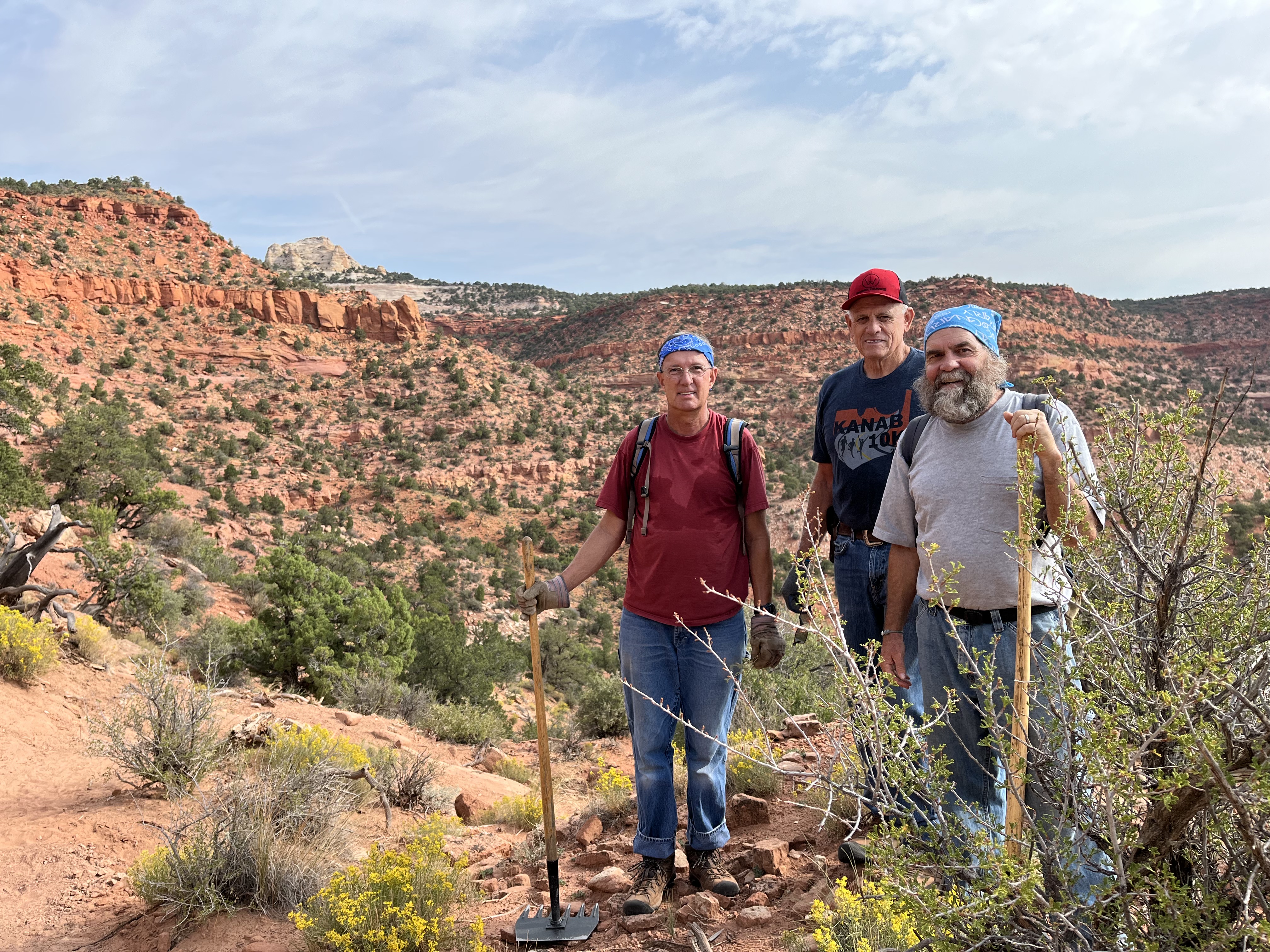 Three volunteers with shovels and rakes stand on a dirt trail outdoors.