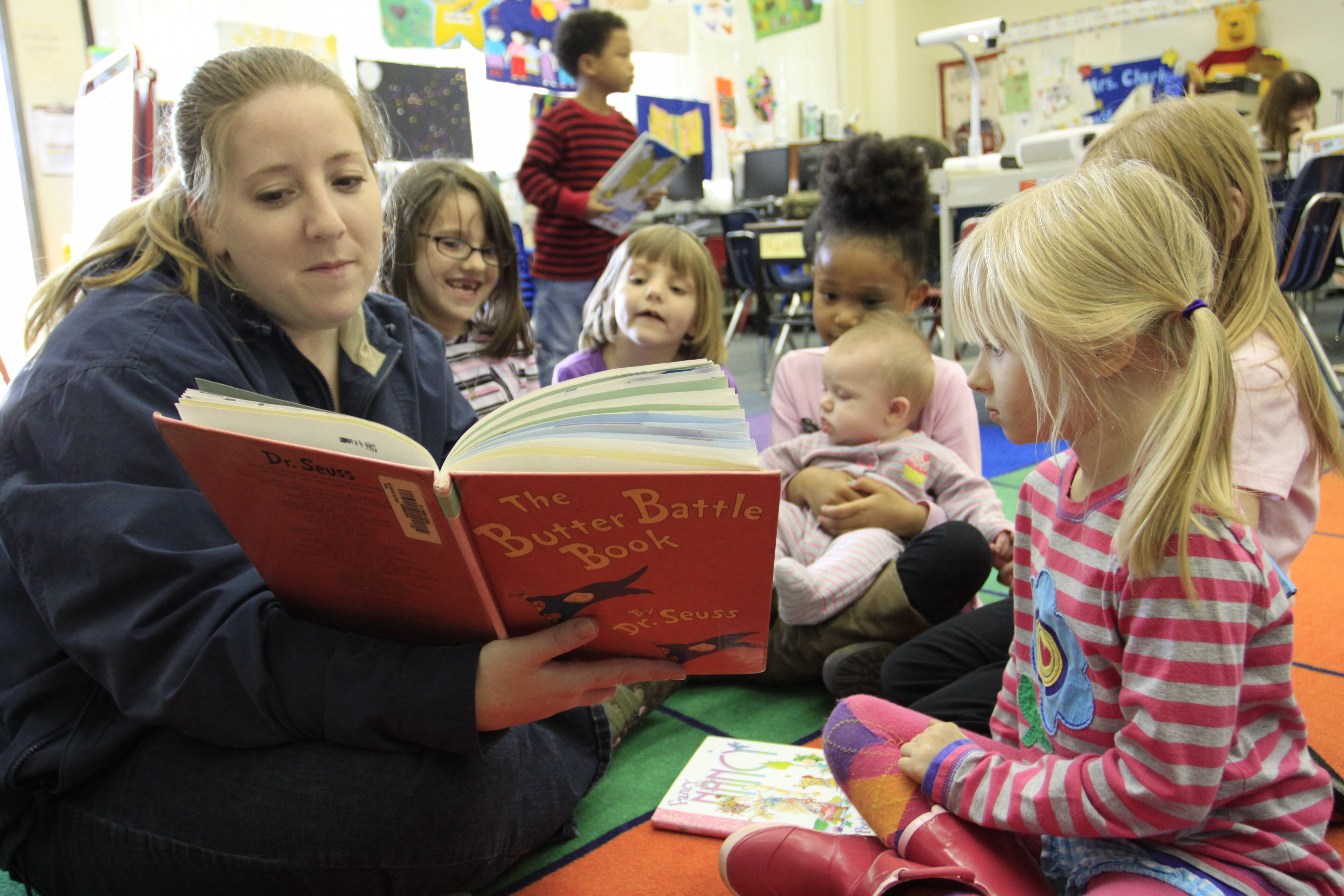 An adult reads a picture book to young children seated on a colorful classroom floor.