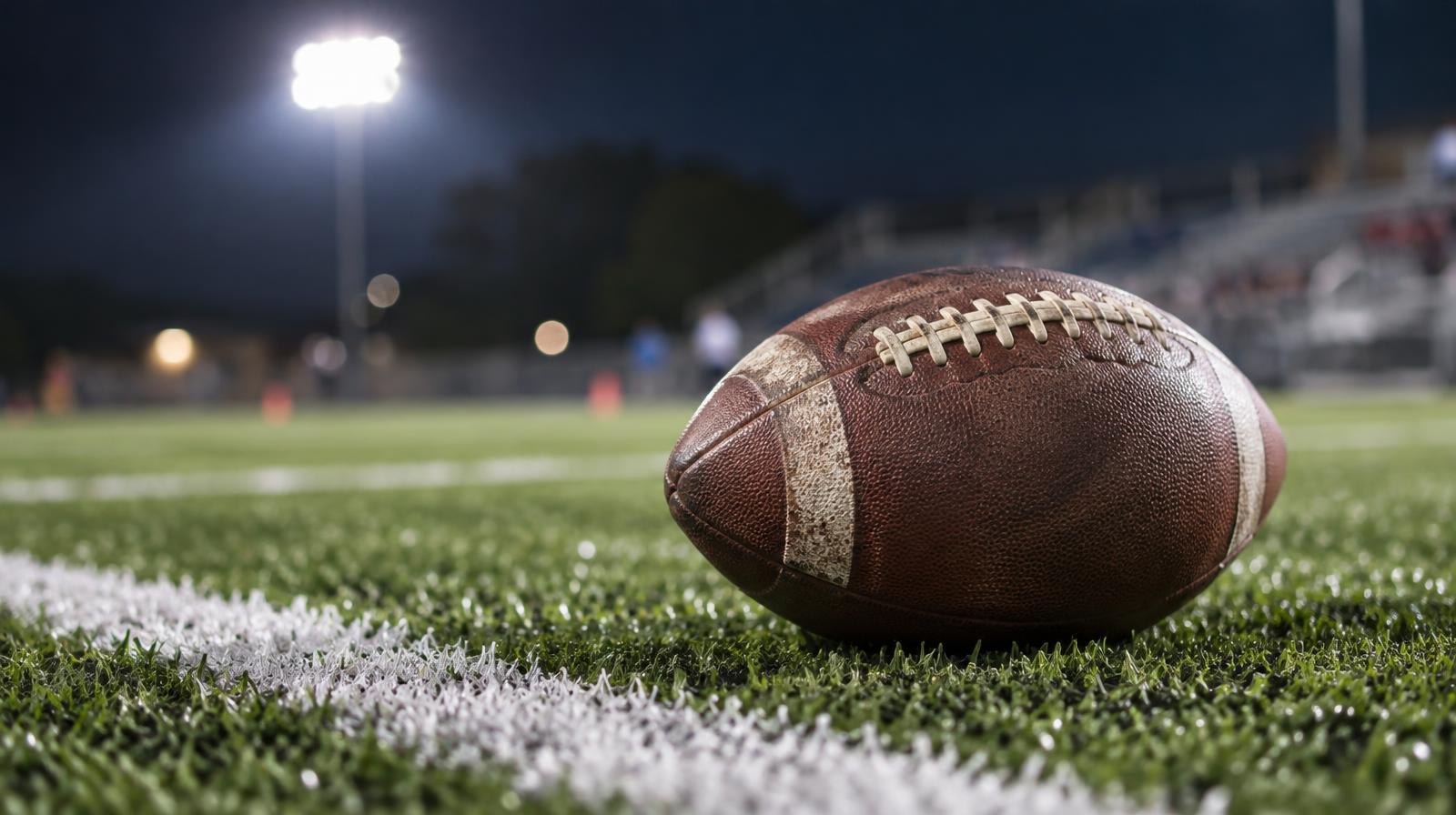 An American football on green artificial turf beside a white yard line under stadium lights.