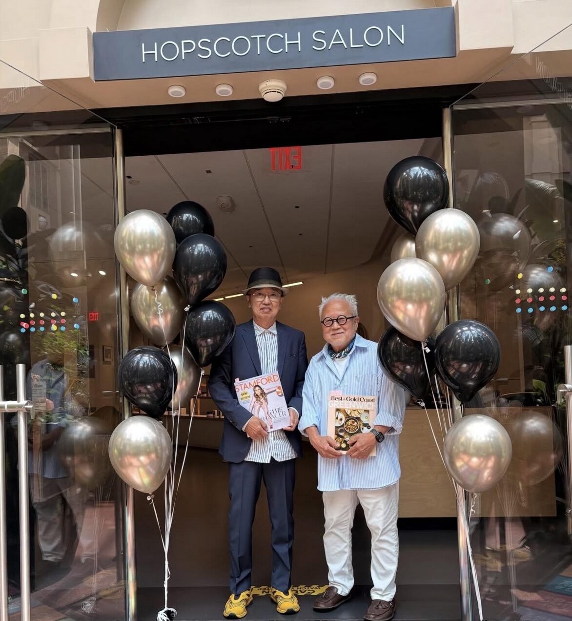 Two men standing in front of Hopscotch Salon entrance flanked by black and silver balloon clusters.