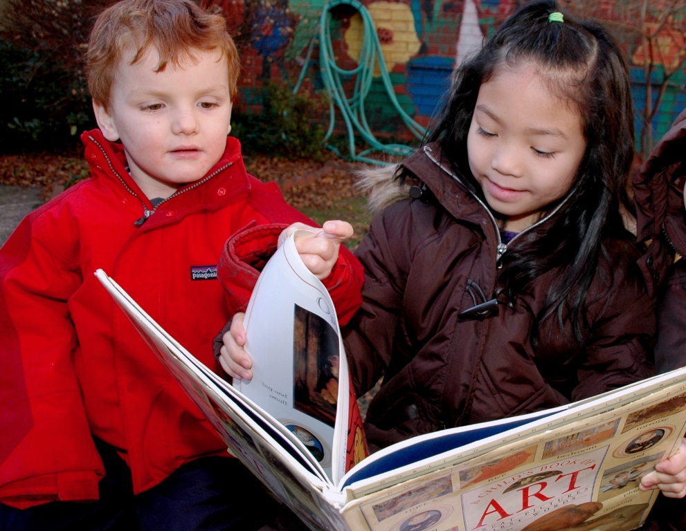 Two young children look at a large illustrated book together outdoors.