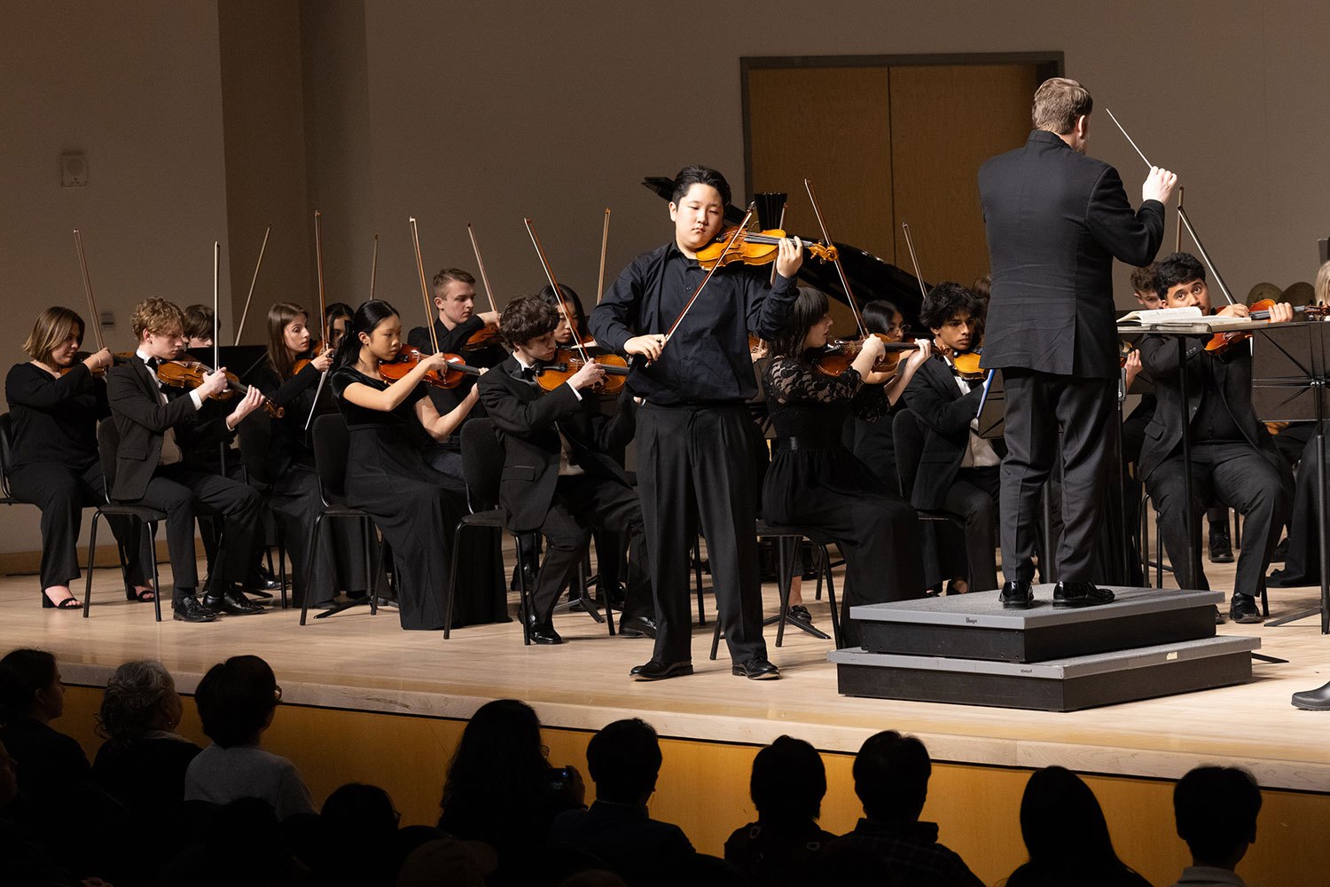 Youth orchestra performs on stage, conductor raises baton before dozens of young musicians in black attire.