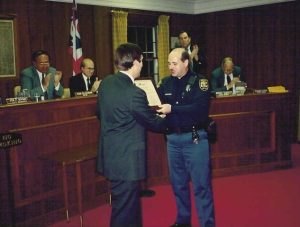 Uniformed police officer shaking hands and receiving a framed document in a government chamber.