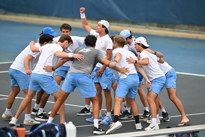 Male tennis players in light blue and white uniforms huddle in celebration on a tennis court.