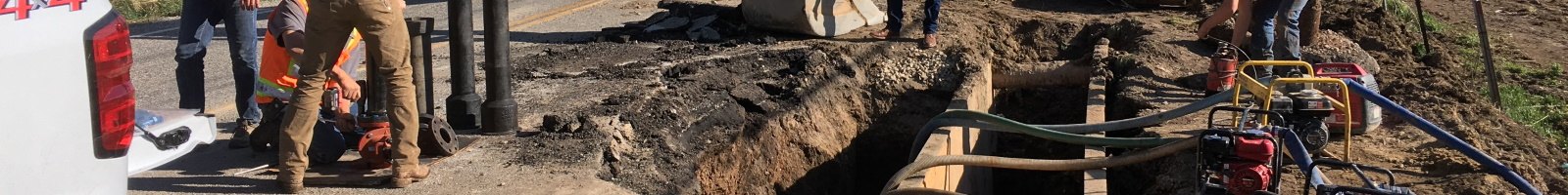 Workers in a trench excavating around exposed underground utility pipes.