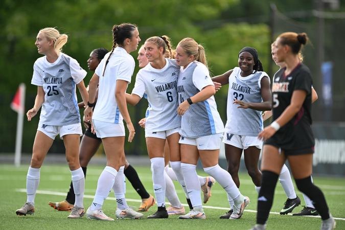 Several soccer players in white and light blue uniforms celebrate together on a grass field.