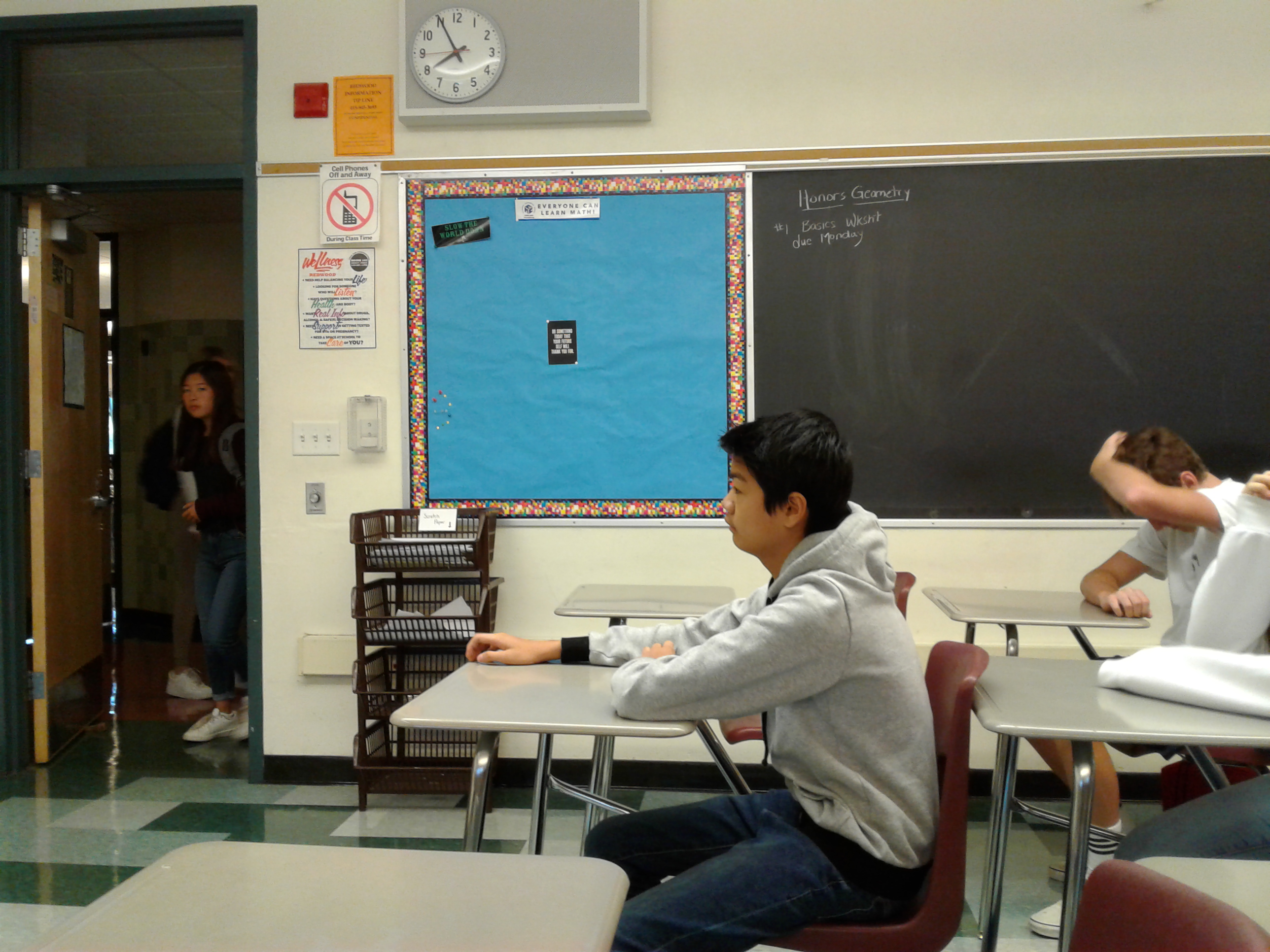 Students seated at desks in a classroom with a chalkboard and bulletin board visible.