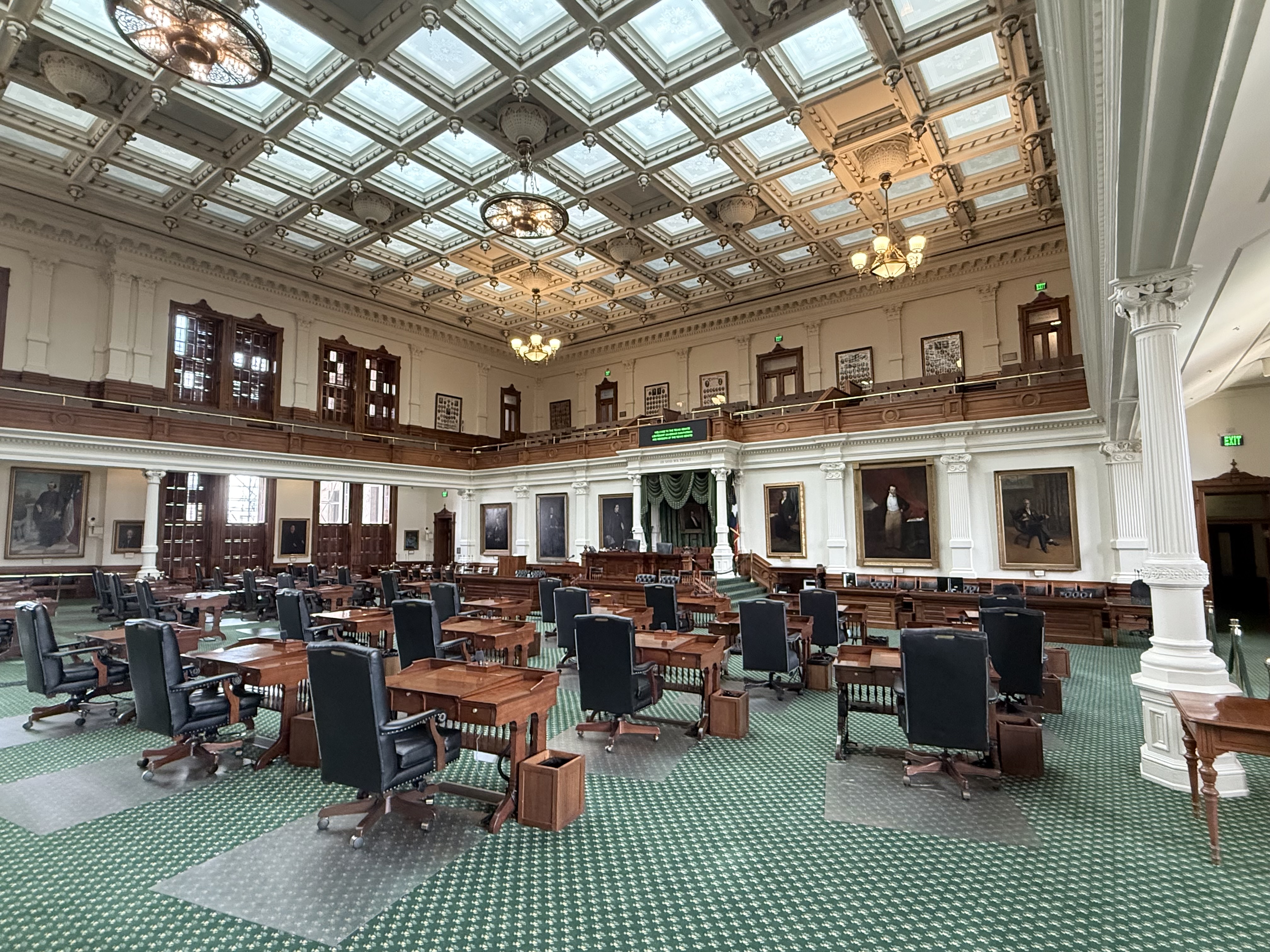 Empty Texas Senate chamber with rows of wooden desks, green carpet, and ornate ceiling.