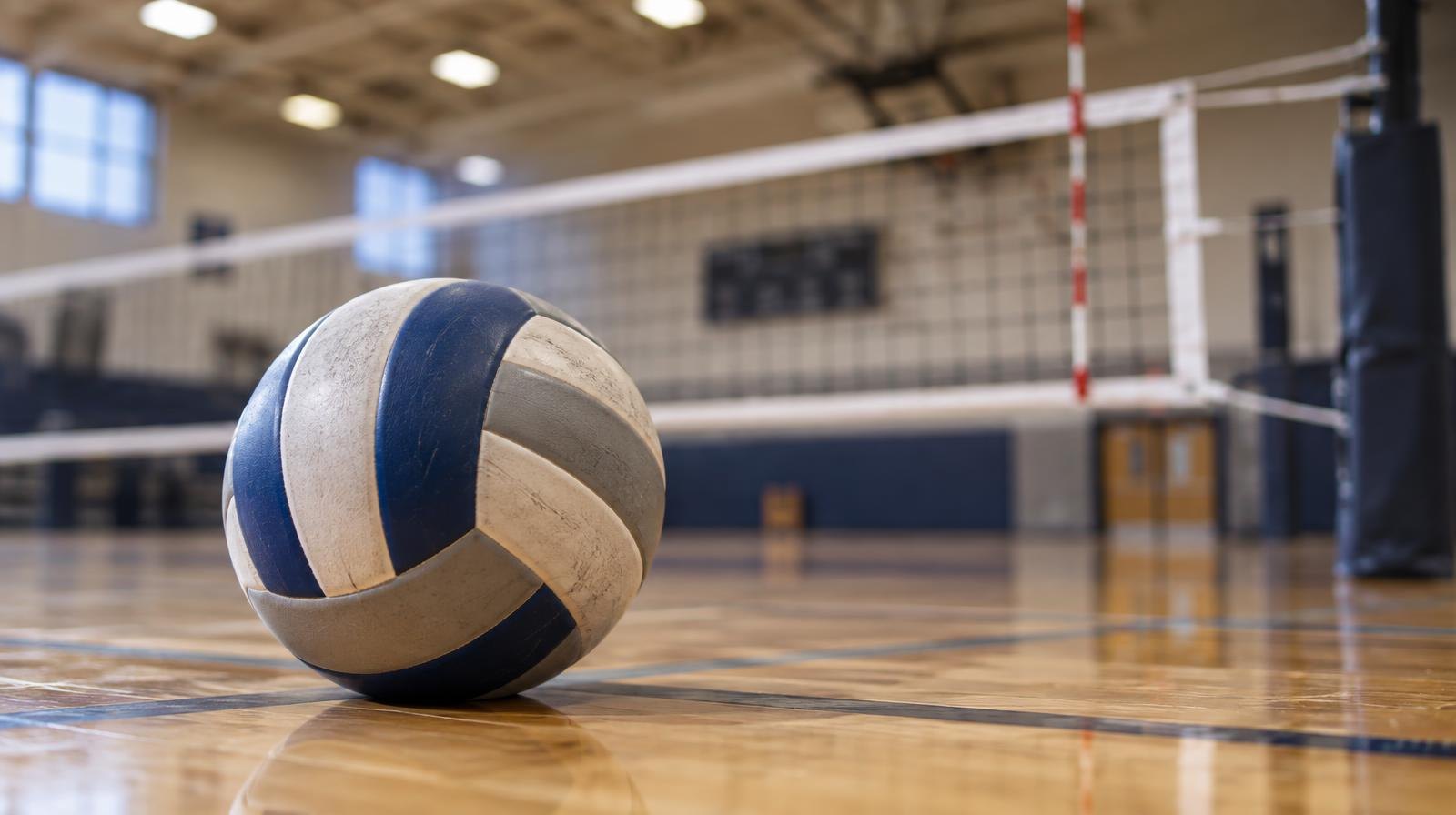 A volleyball resting on a hardwood gym floor near a volleyball net.
