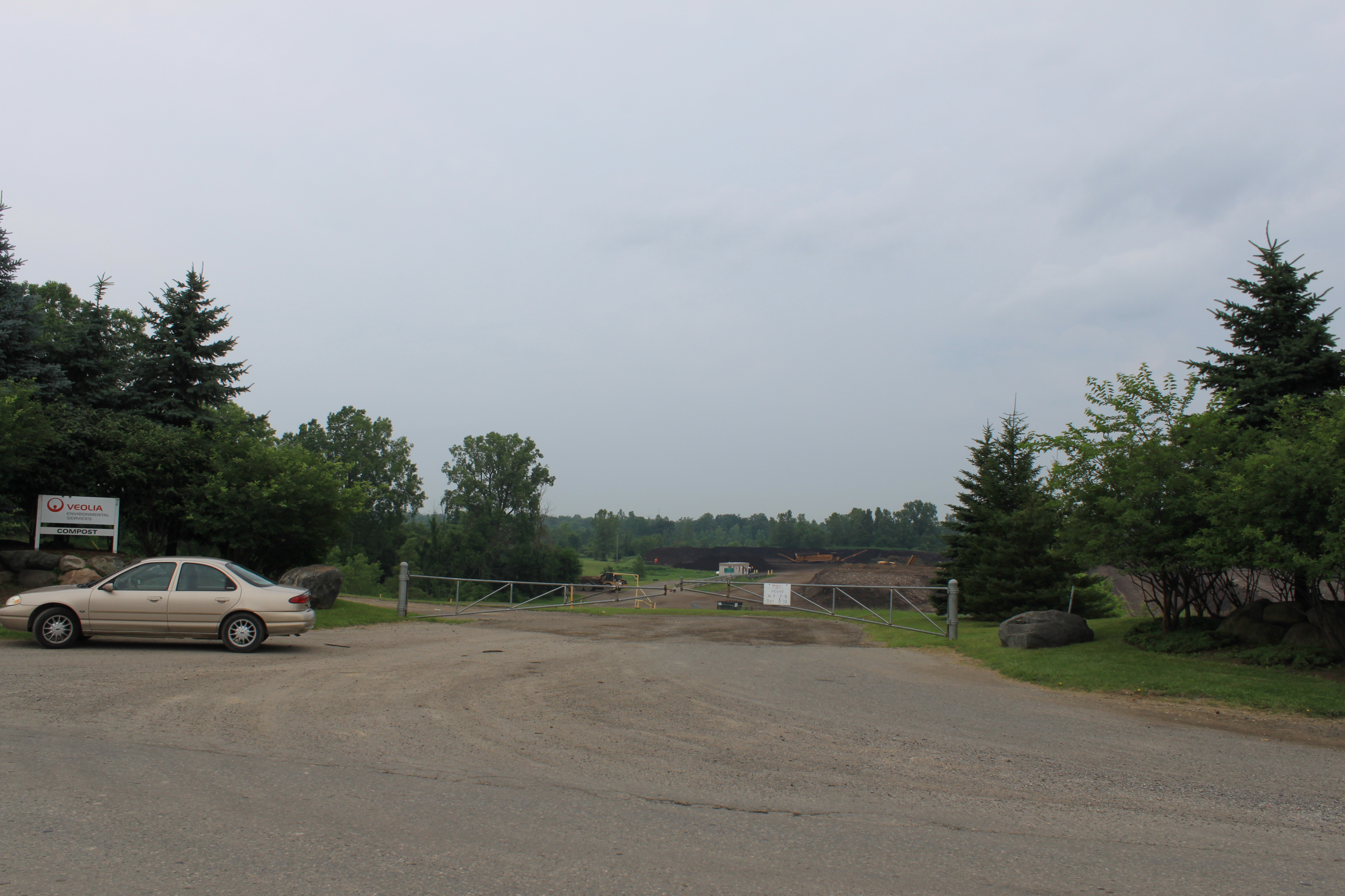 Gated entrance to a landfill facility flanked by evergreen trees under an overcast sky.