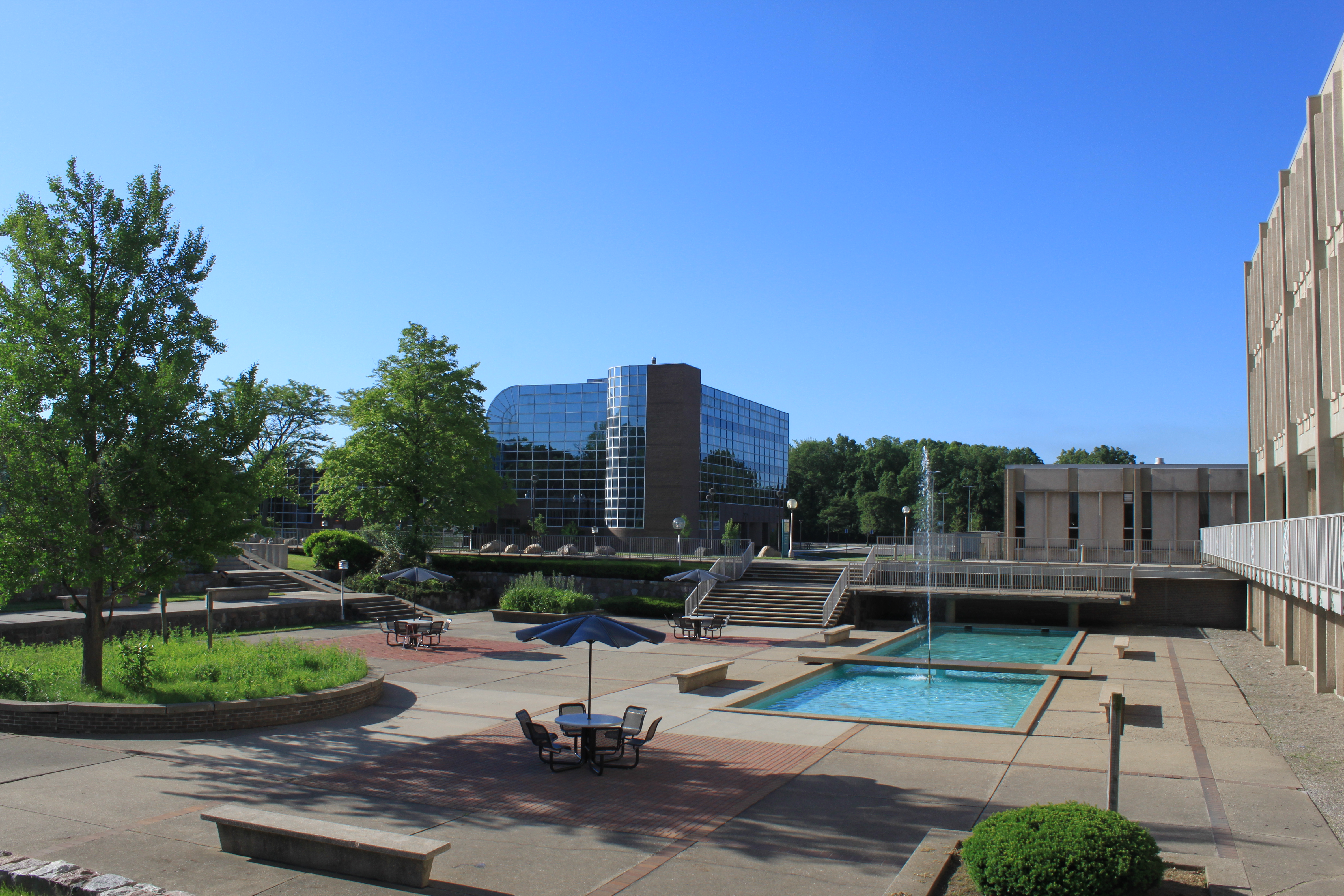 Outdoor college campus plaza with a fountain, umbrella tables, and a glass-facade building.