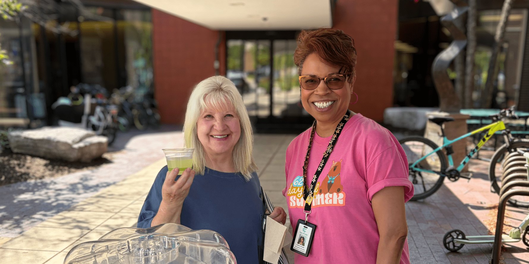 Two smiling women outdoors near a brick building, one holding a tray of cups.