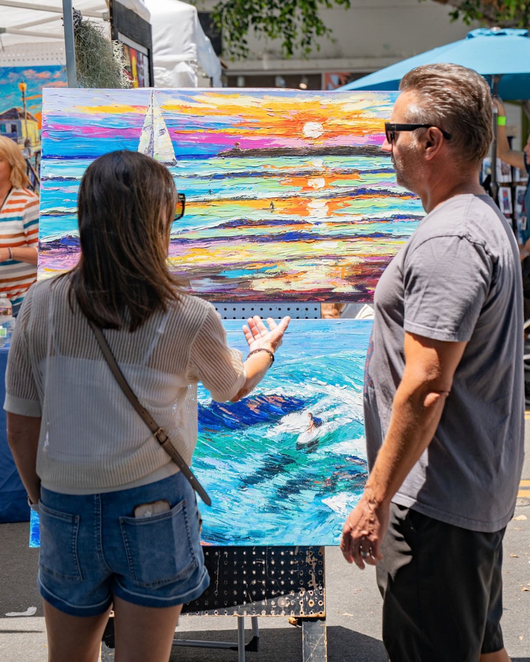 Two people stand before a large colorful ocean painting at an outdoor art fair.