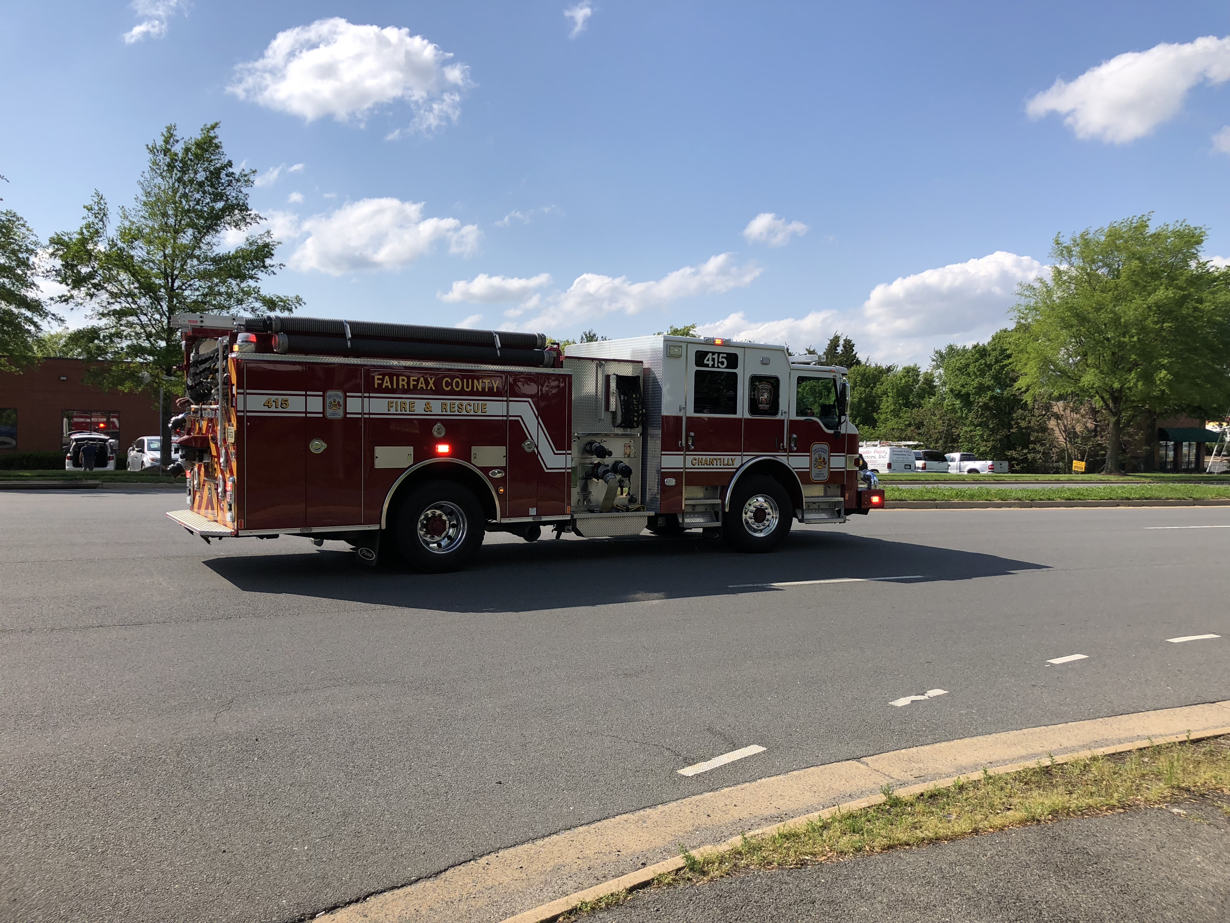 A red and white Fairfax County Fire and Rescue engine driving on a road with emergency lights on.