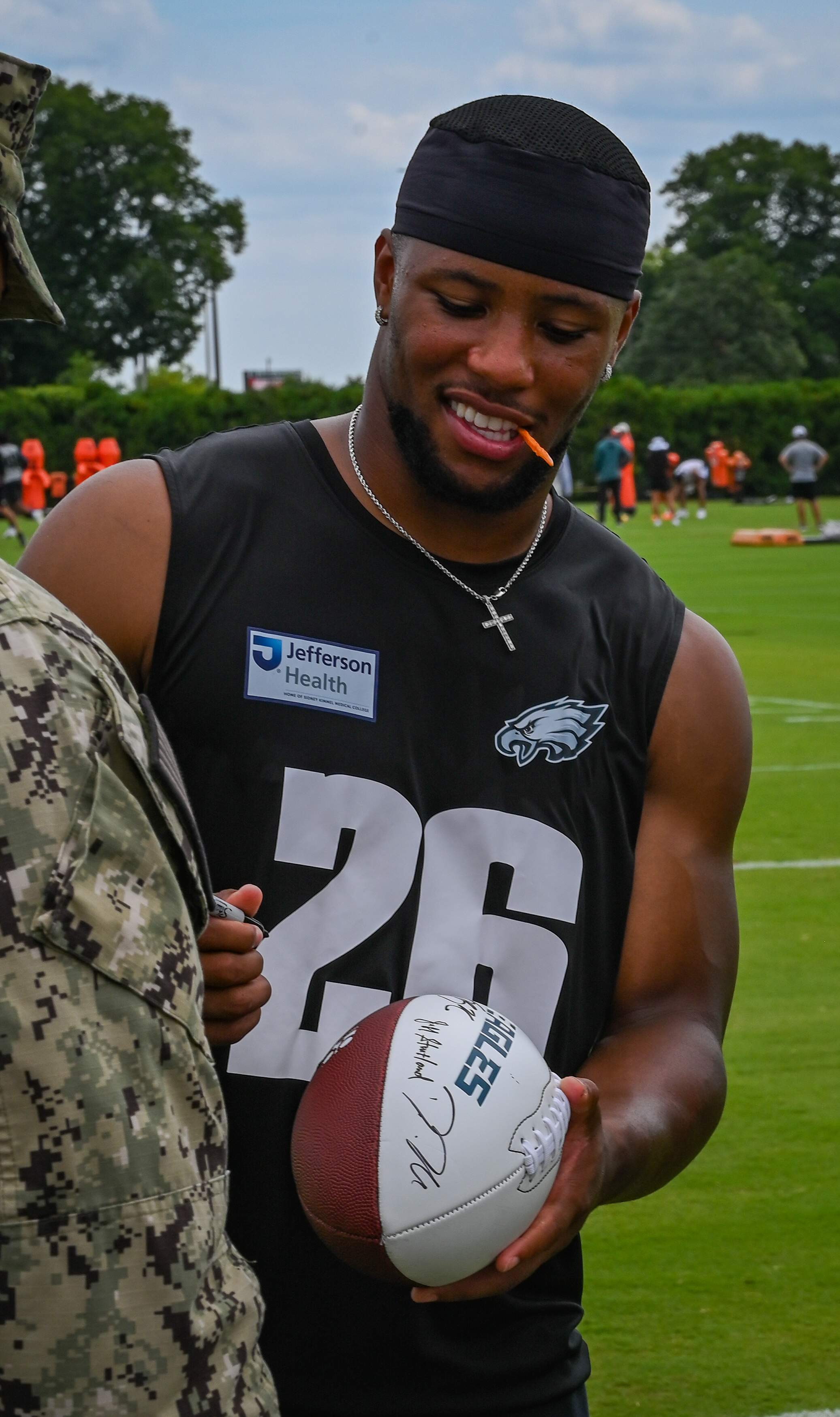 Eagles player in number 26 jersey signing a football at an outdoor practice field.