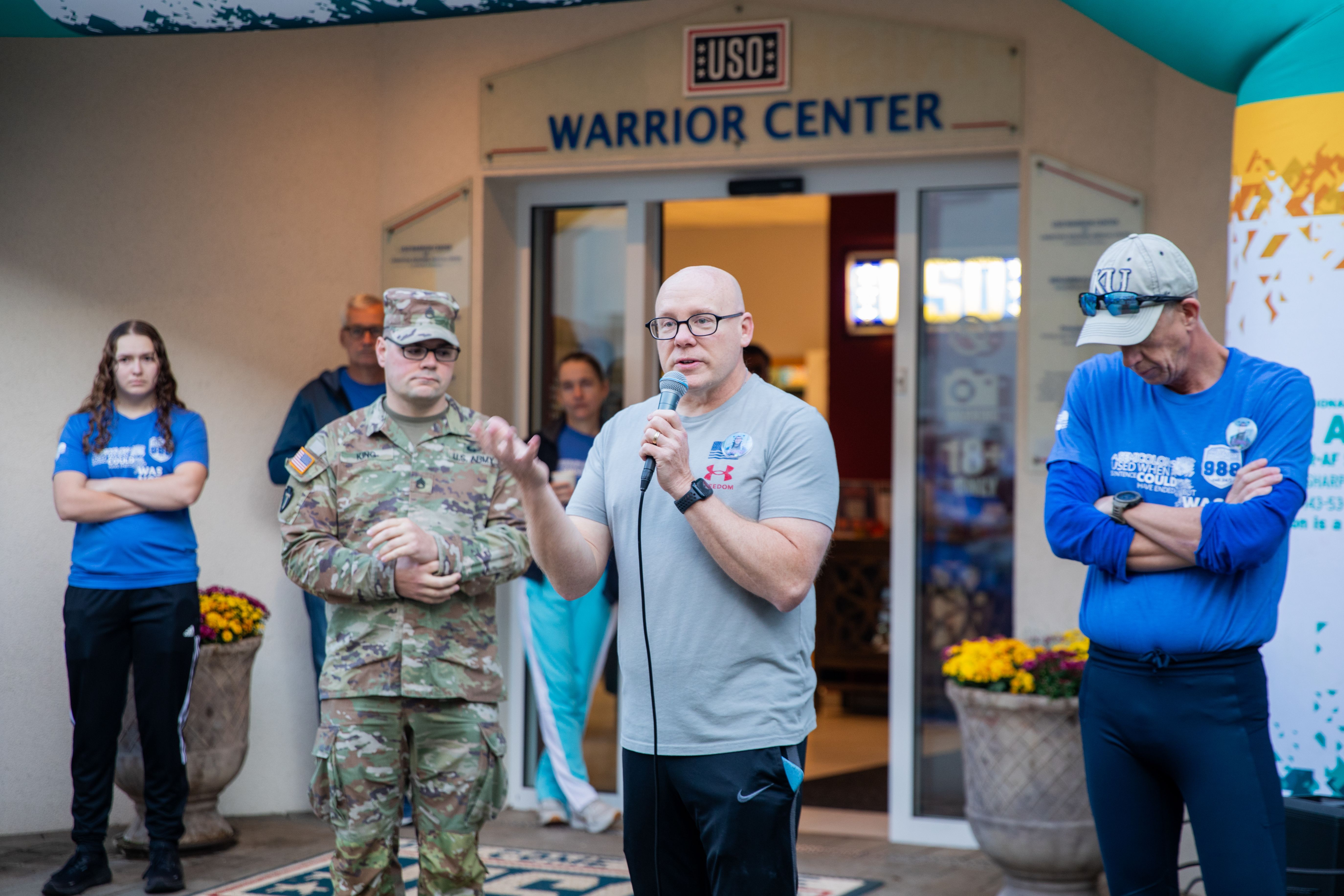 Man speaks at microphone at an outdoor suicide prevention awareness event near a military building.