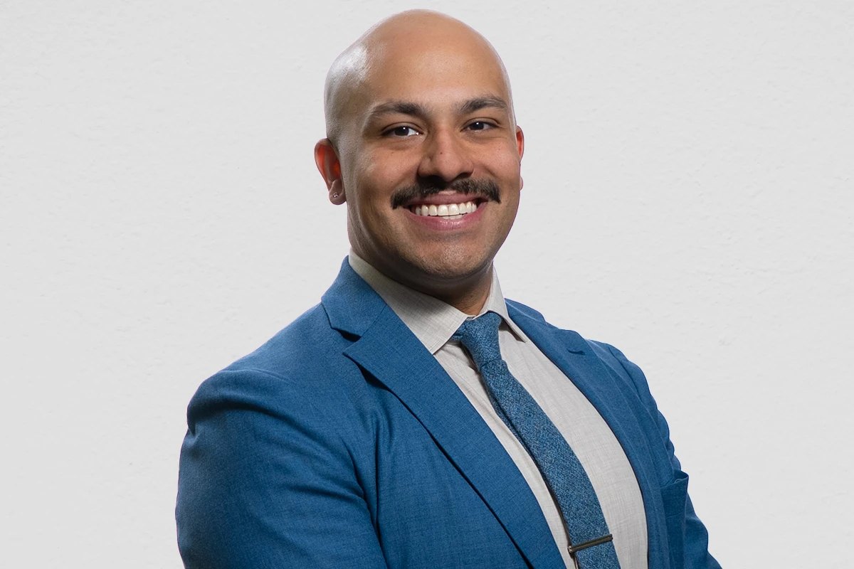 A bald man with a mustache in a blue suit and gray tie smiling in a professional headshot.