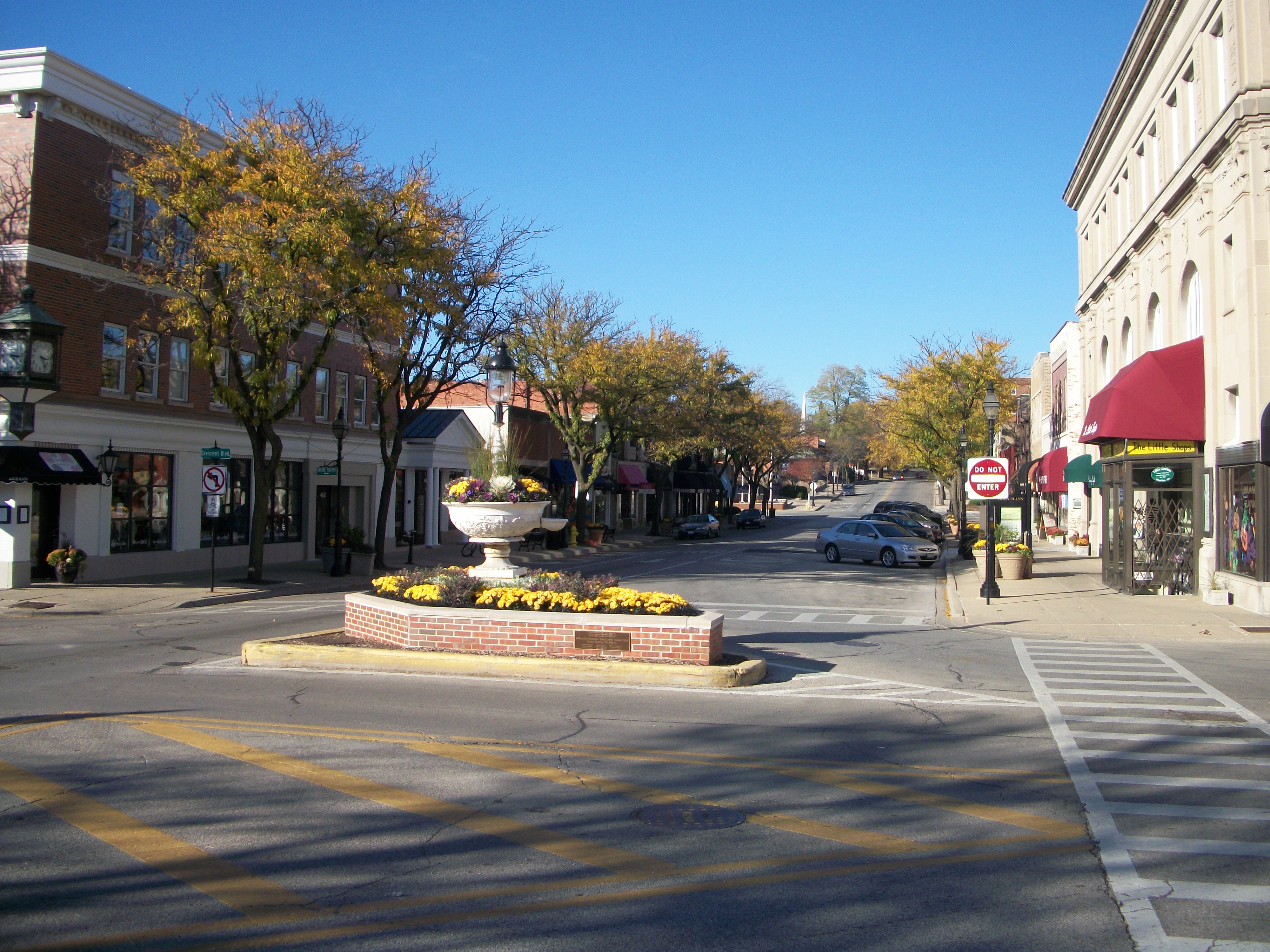 Downtown street intersection with a brick planter, autumn trees, and brick storefronts.