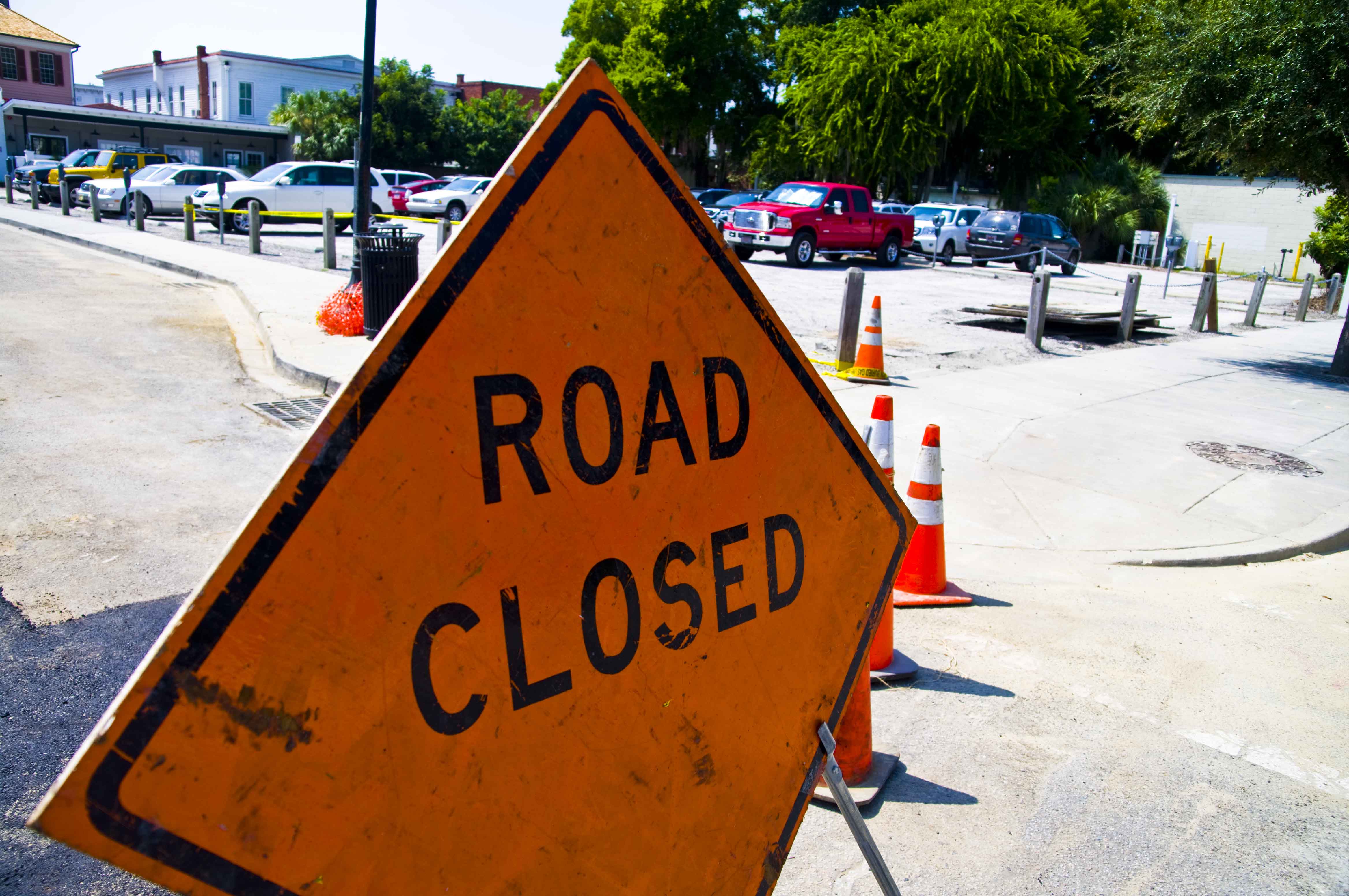 Orange diamond-shaped Road Closed sign on a stand with traffic cones behind it.