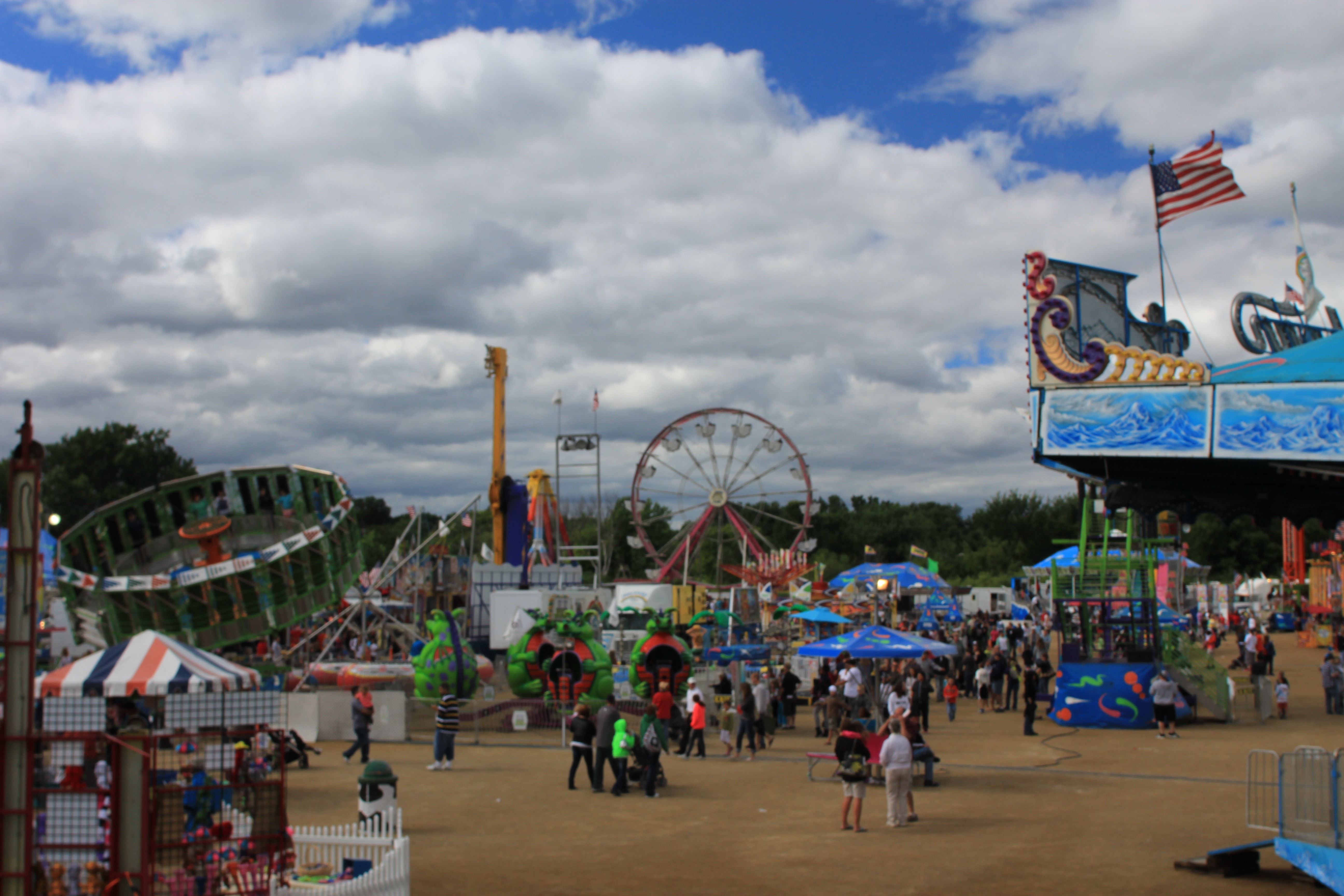 Carnival rides and crowds line the midway at a county fair under a partly cloudy sky.