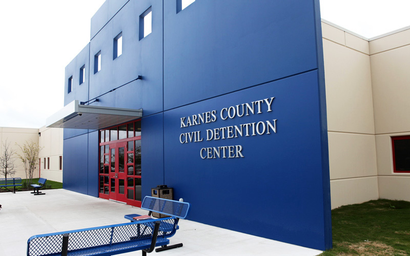 Blue-paneled detention facility building with red entry doors and outdoor benches.