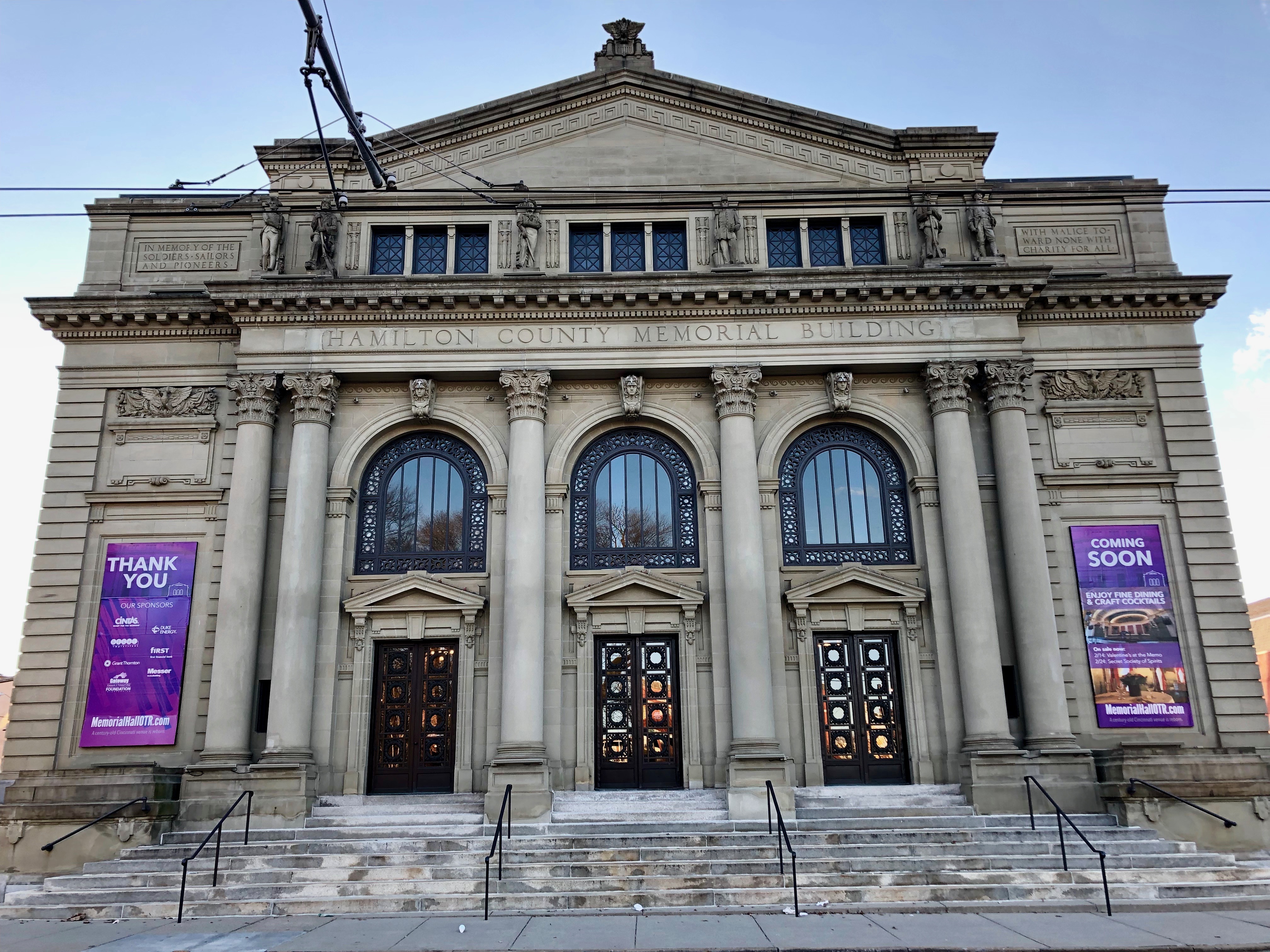 Neoclassical stone building with columns and arched windows, purple banners flanking the entrance steps.