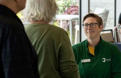 Staff member in green uniform smiling while speaking with two visitors indoors.