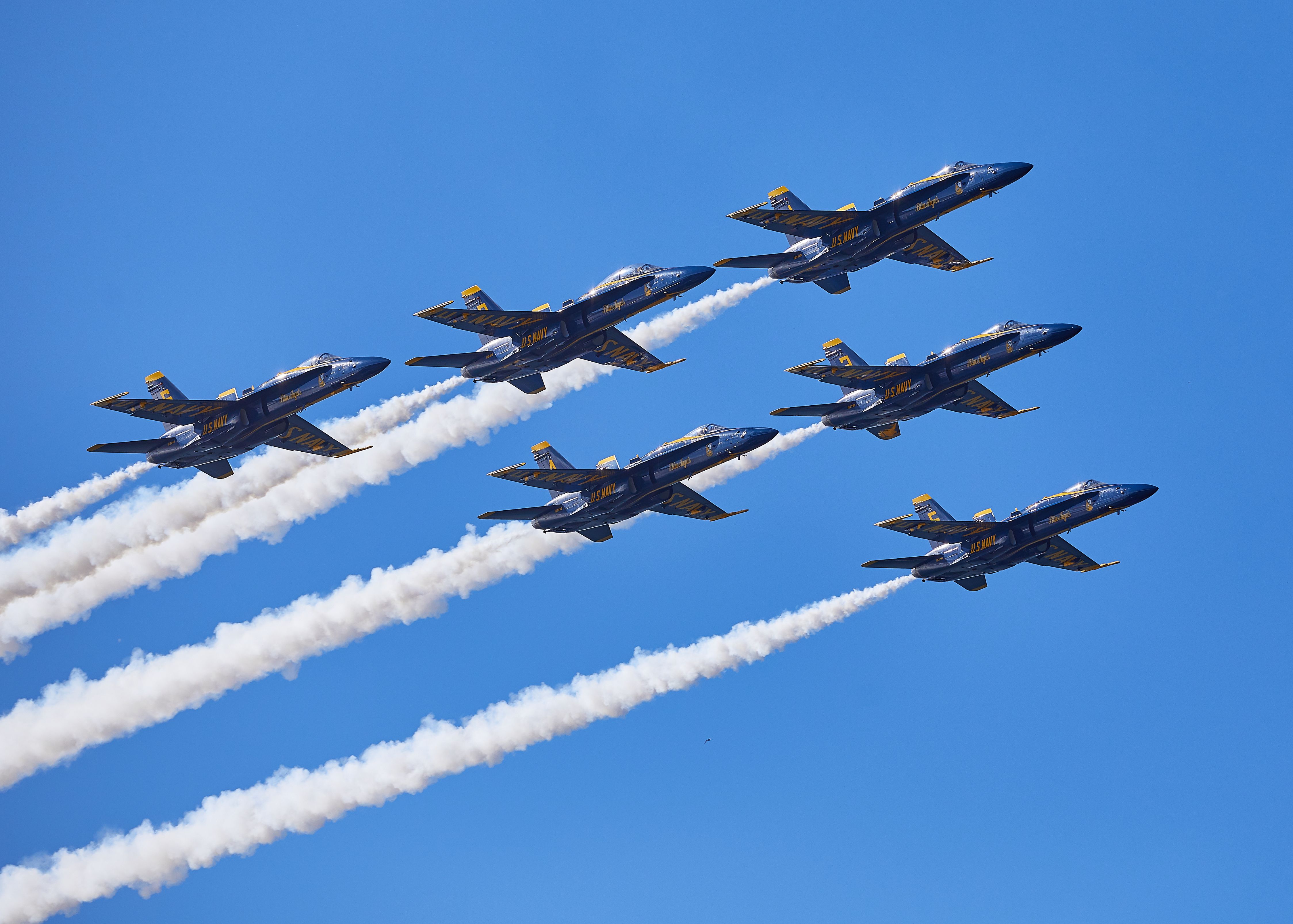Six blue and gold military jets in tight delta formation trailing white smoke against a blue sky.