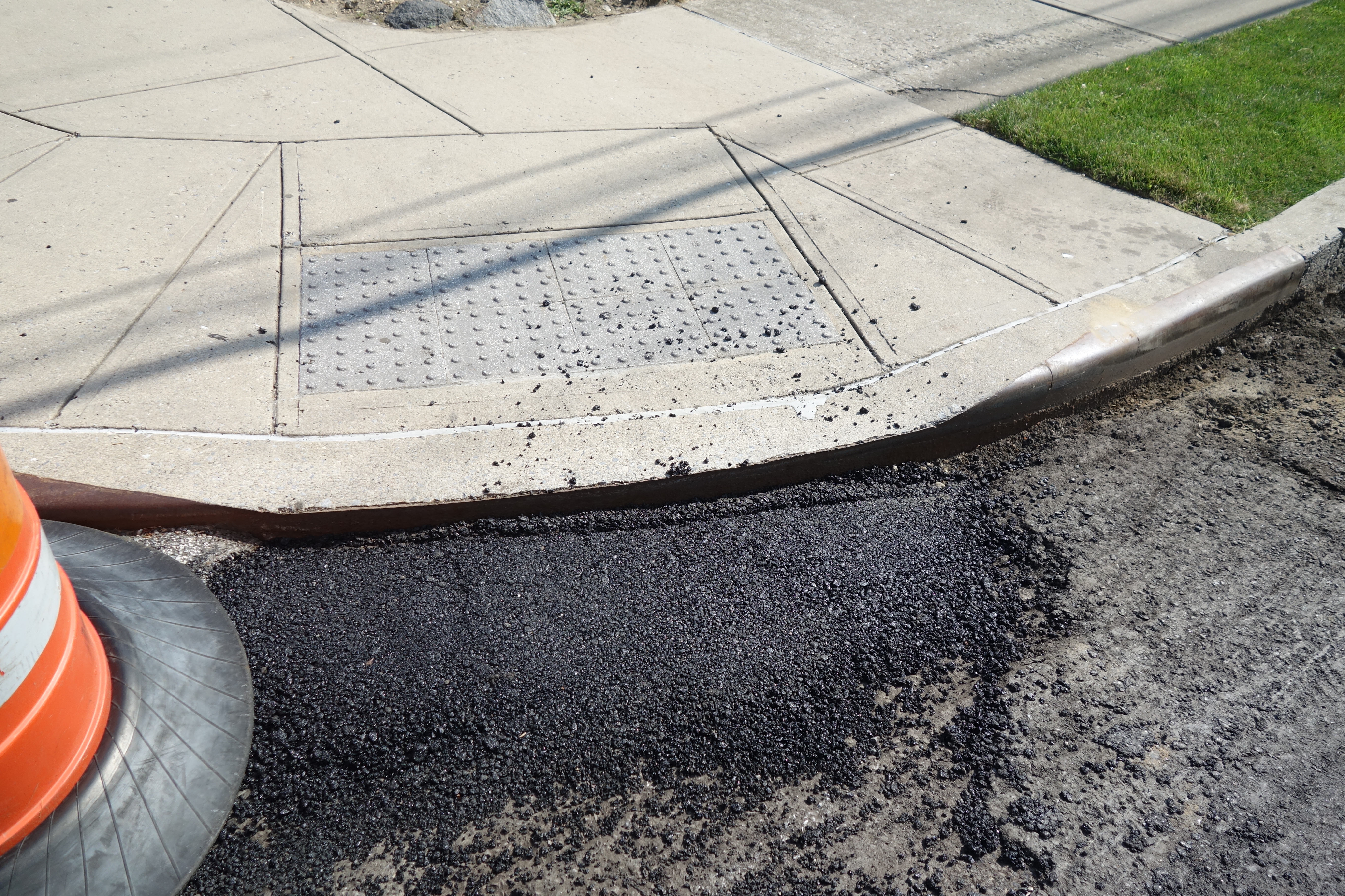 Freshly laid asphalt at a curb cut with an orange traffic cone nearby.