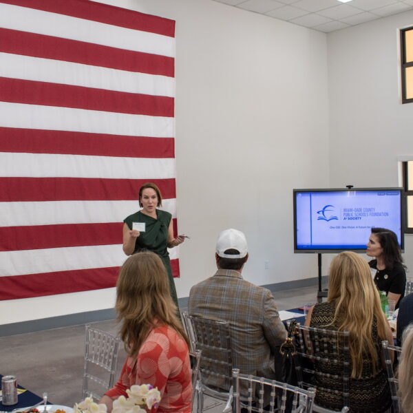 A woman in green addresses seated attendees at an M-DCPS Foundation event, with an American flag behind her.