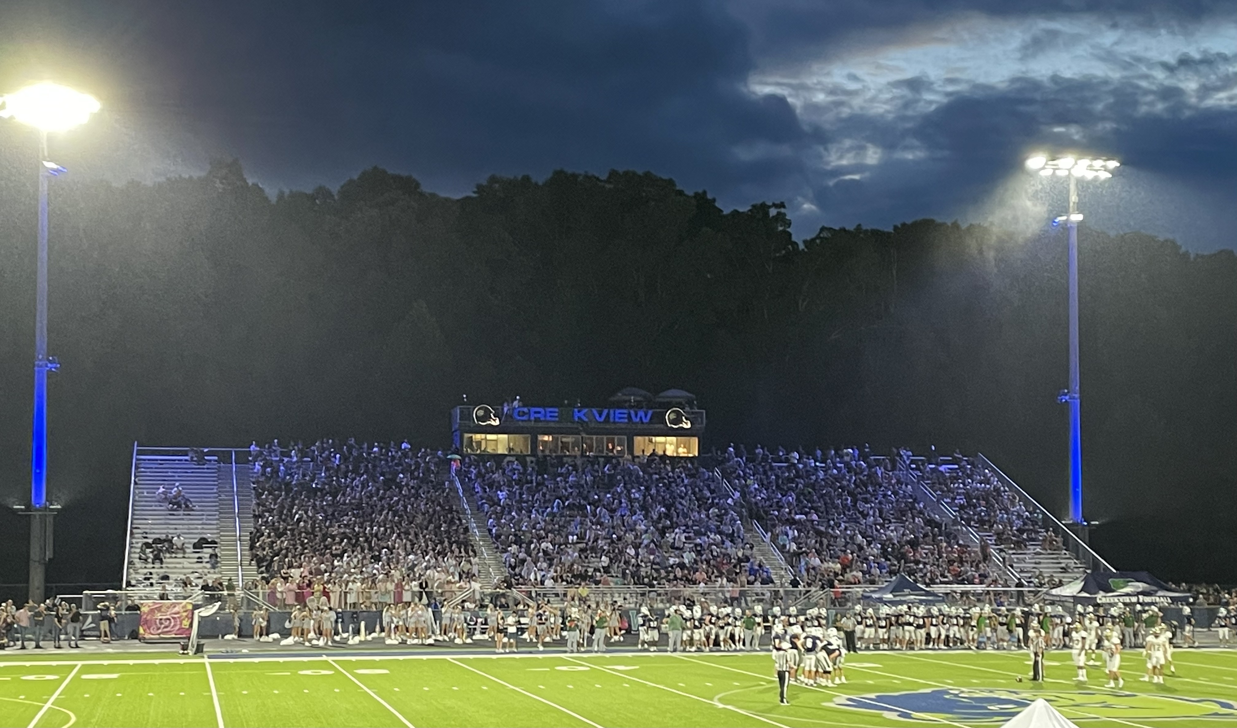 Packed high school football stadium at night with bright lights and a large scoreboard.