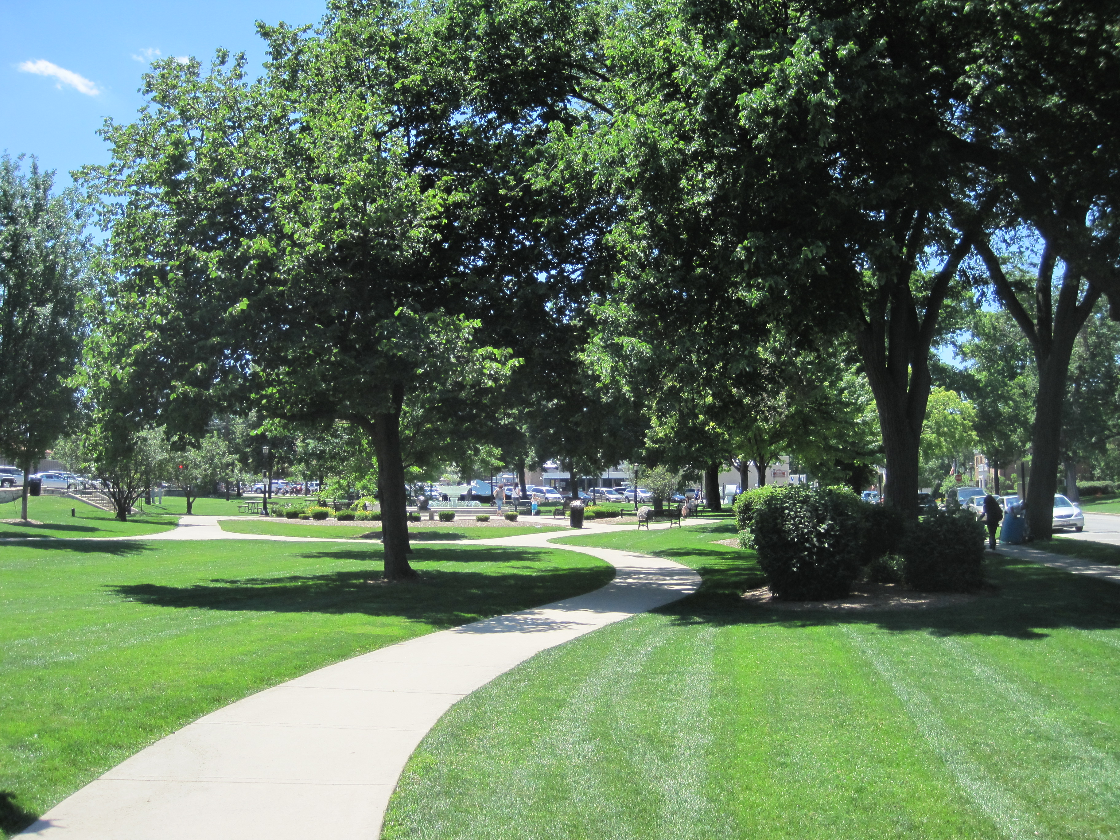 Tree-lined park with a curved concrete walkway and manicured green lawn on a sunny day.