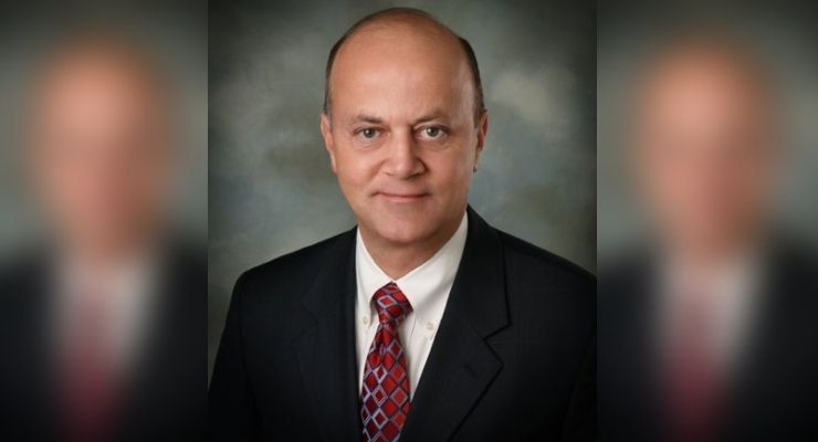 Professional headshot of a bald man in a dark suit, white shirt, and red patterned tie.