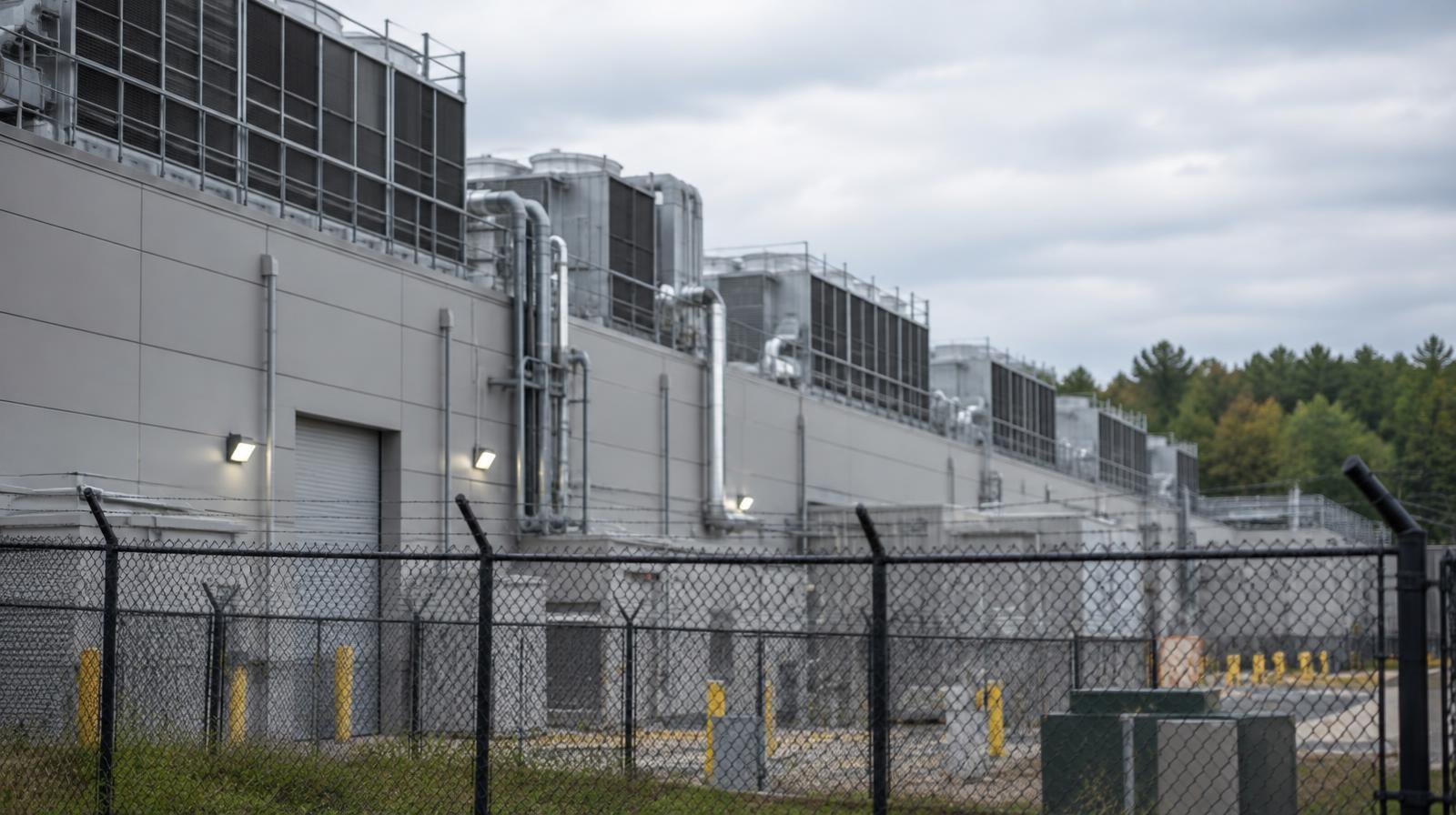 Large industrial building with rooftop cooling units behind a chain-link security fence.