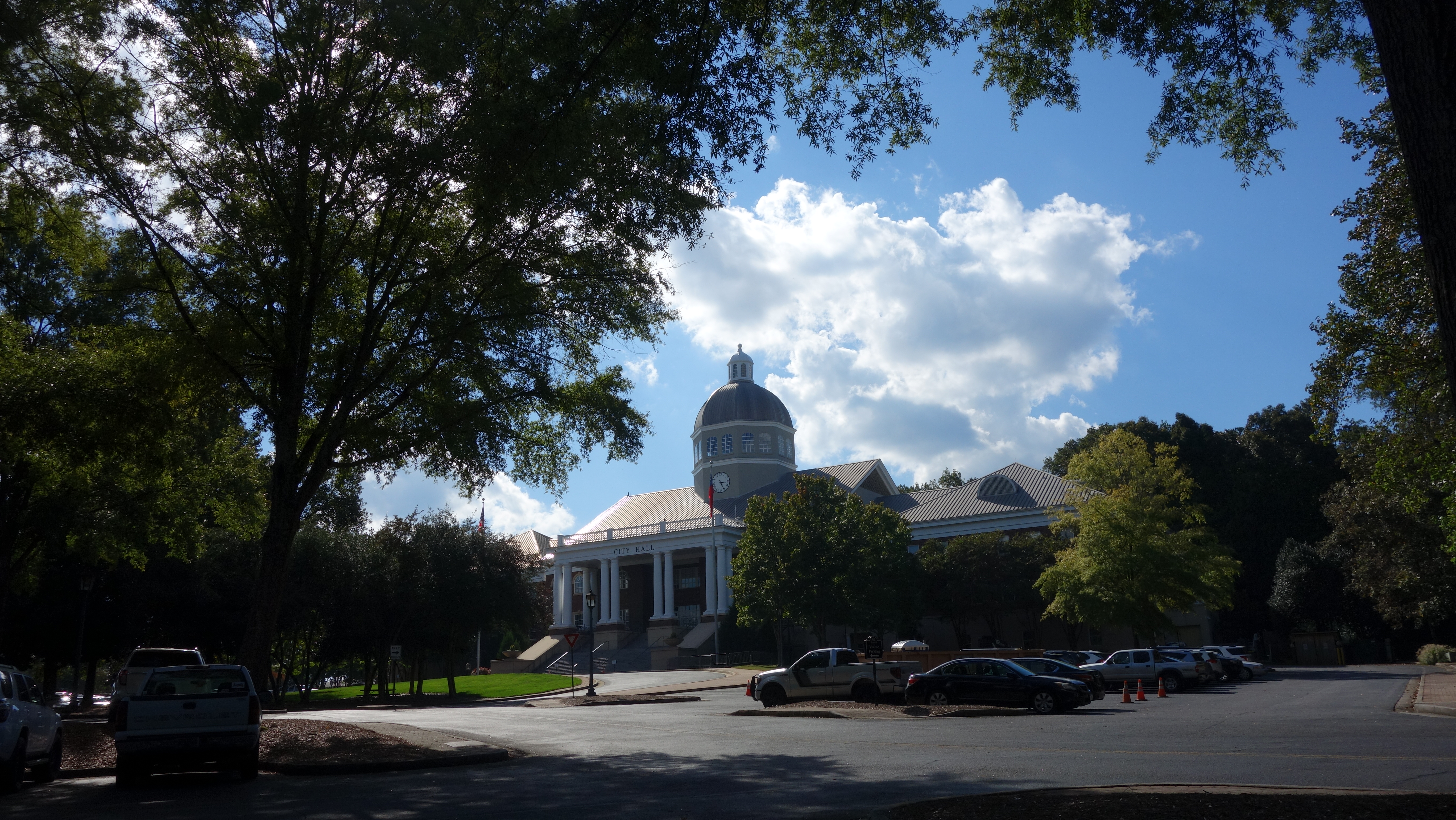 Domed brick government building seen through mature trees with parked cars.