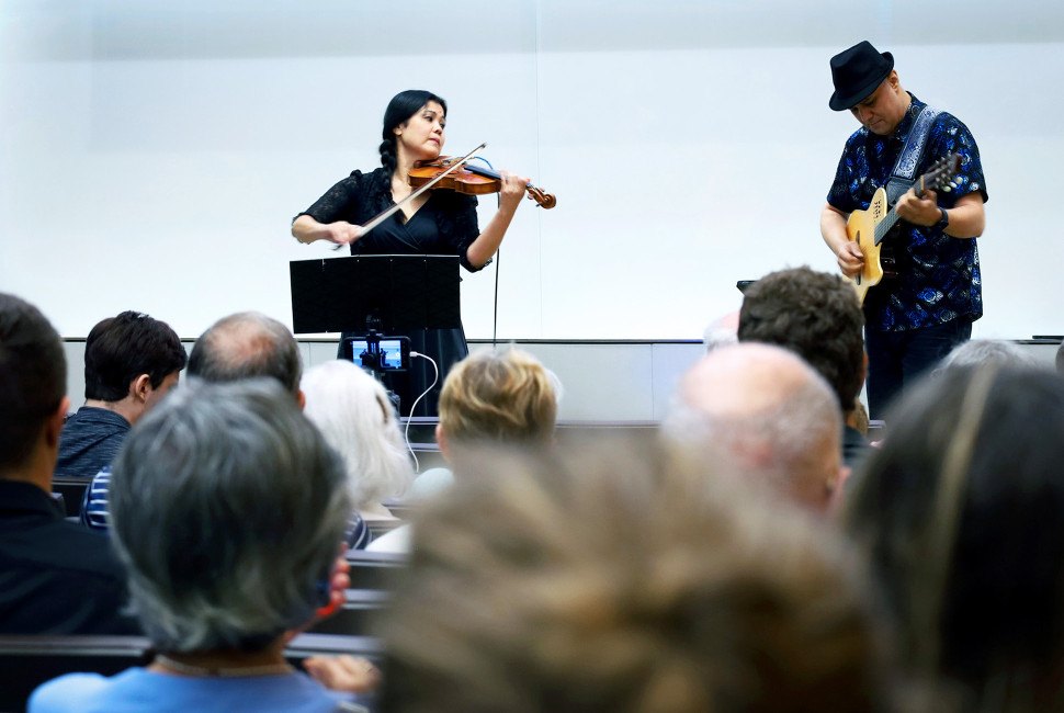A woman plays violin and a man plays guitar on a small stage before a seated audience.