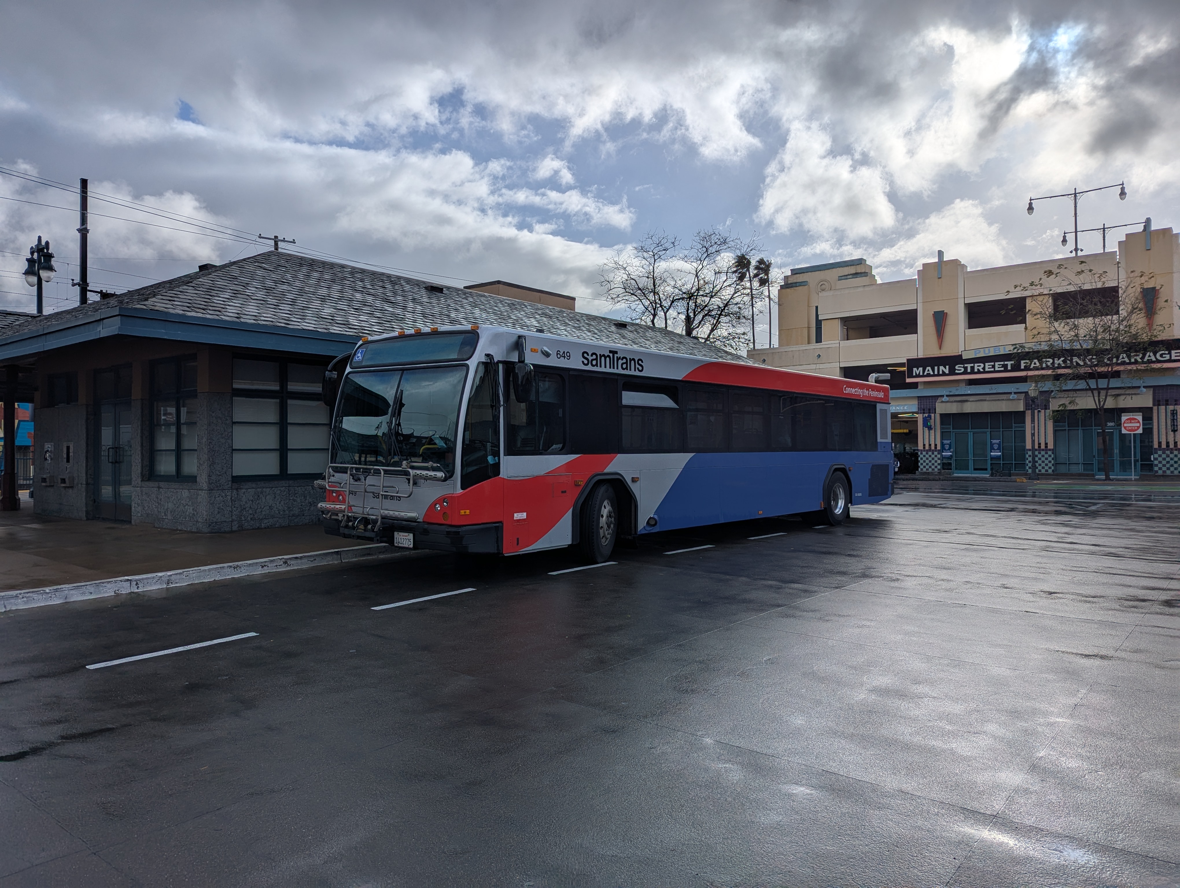 A red, white, and blue SamTrans bus parked at a covered station on a wet day.
