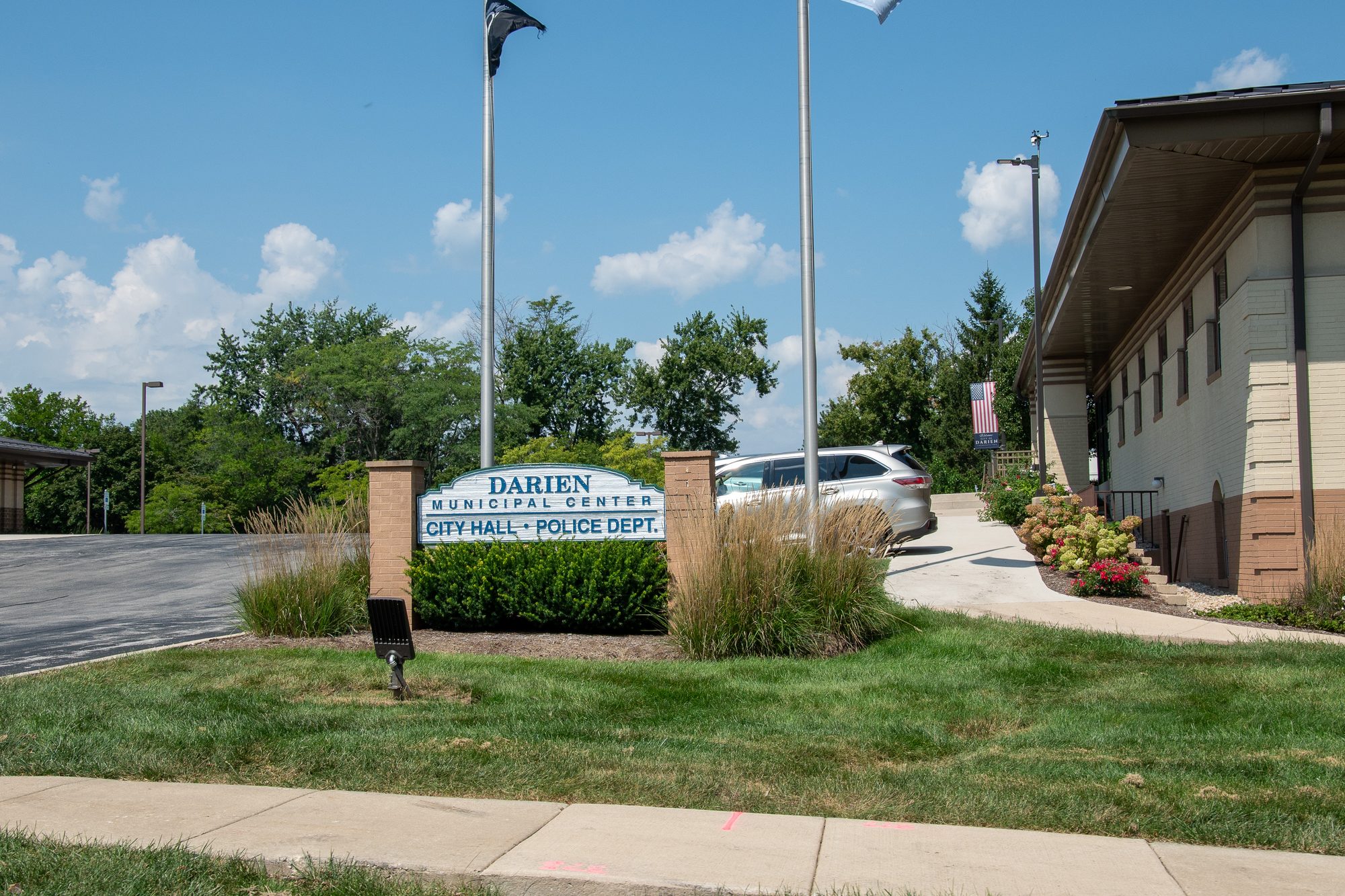 Exterior of a low-rise municipal center with flagpoles and a parked white SUV.