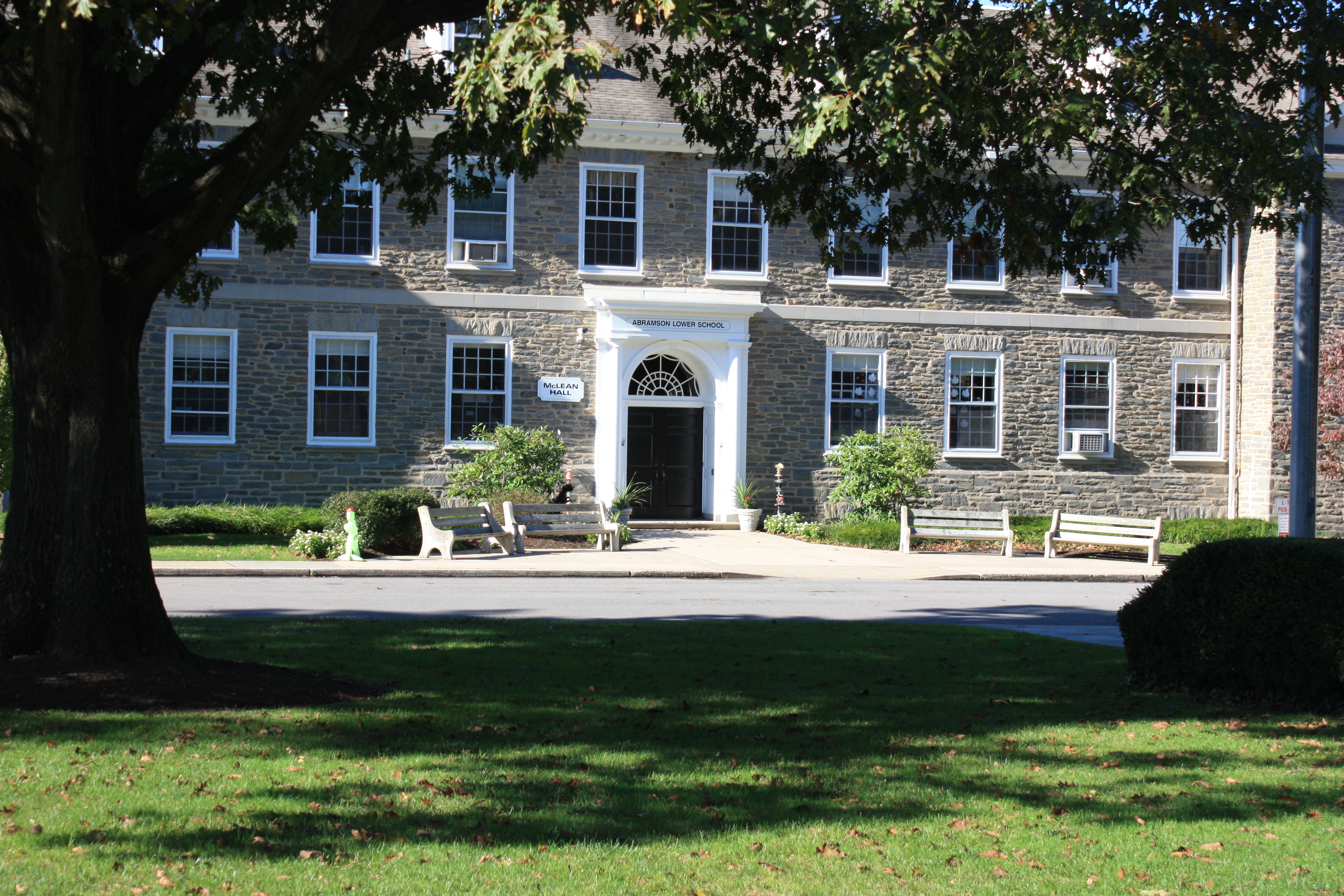 Stone lower school building with white-trimmed windows, benches, and mature trees.