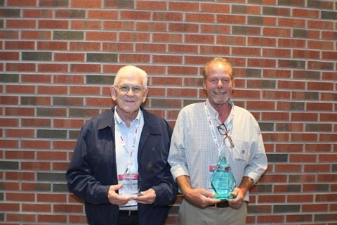 Two men stand before a brick wall, each holding a glass award trophy.