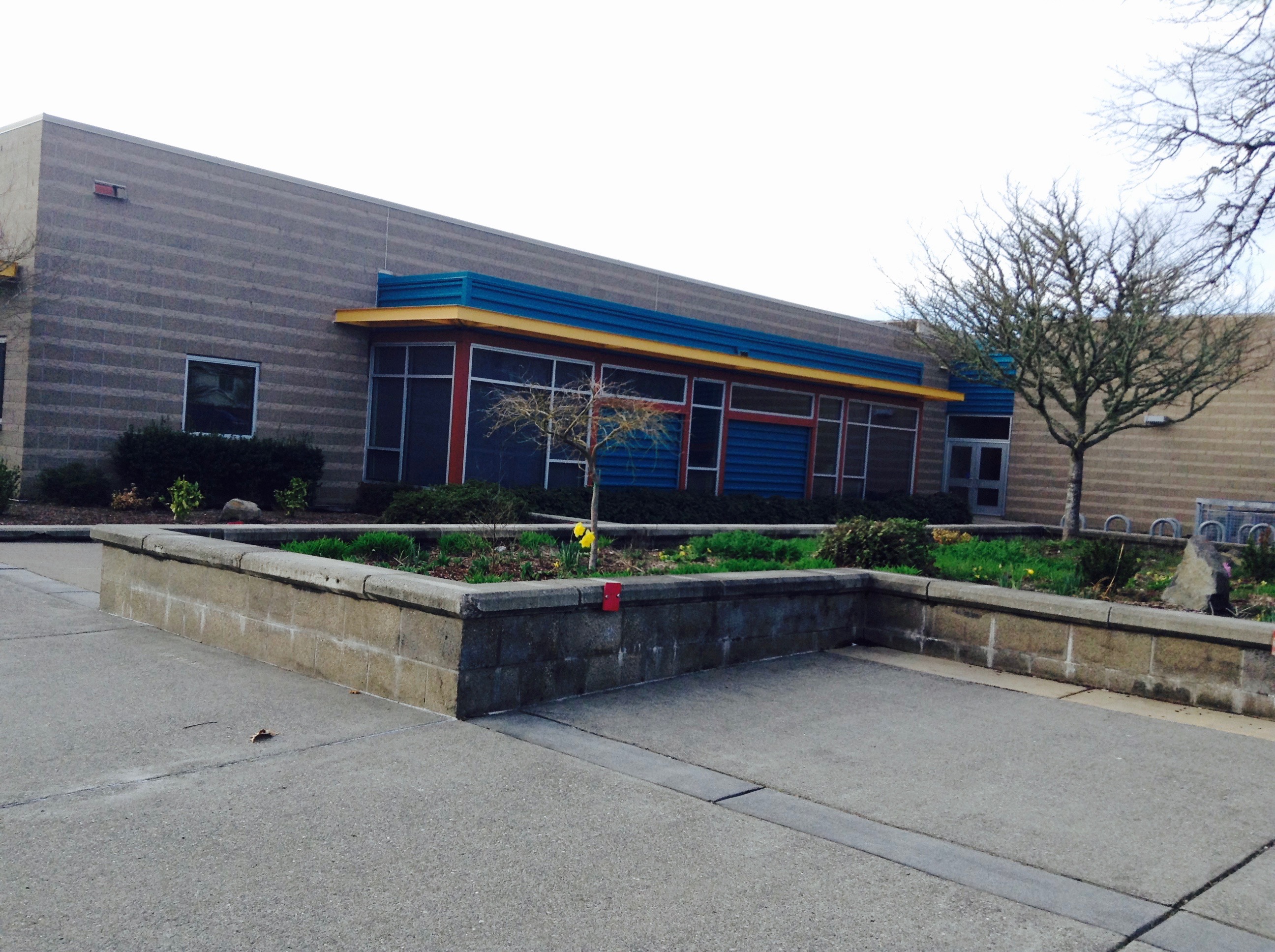 Single-story school building with blue and yellow trim and a raised stone planter in front.