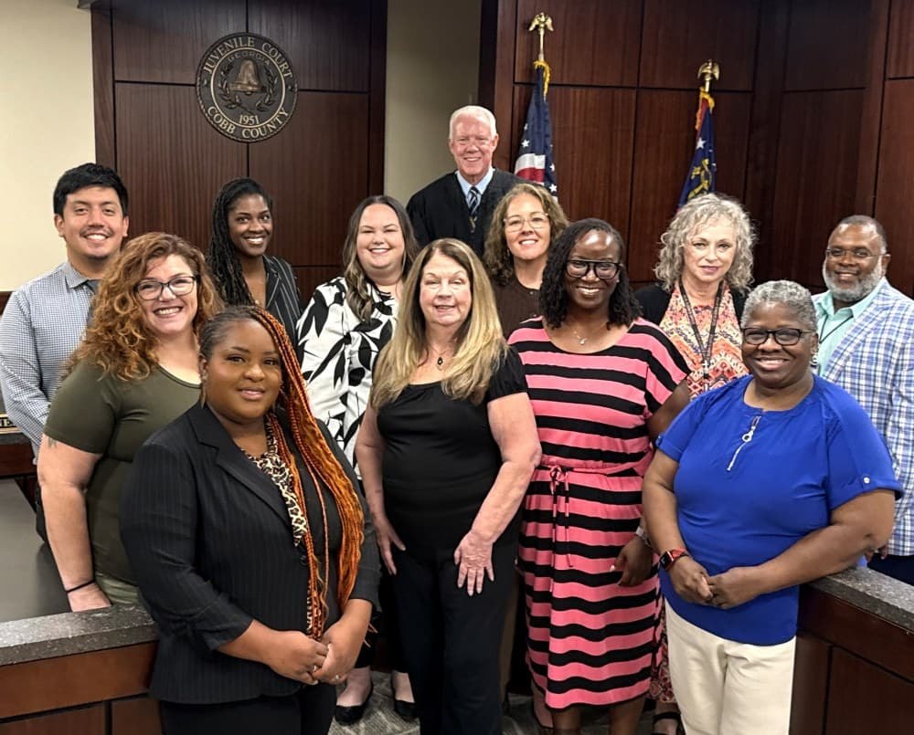 Eleven people pose together in a wood-paneled courtroom with an American flag and court seal.