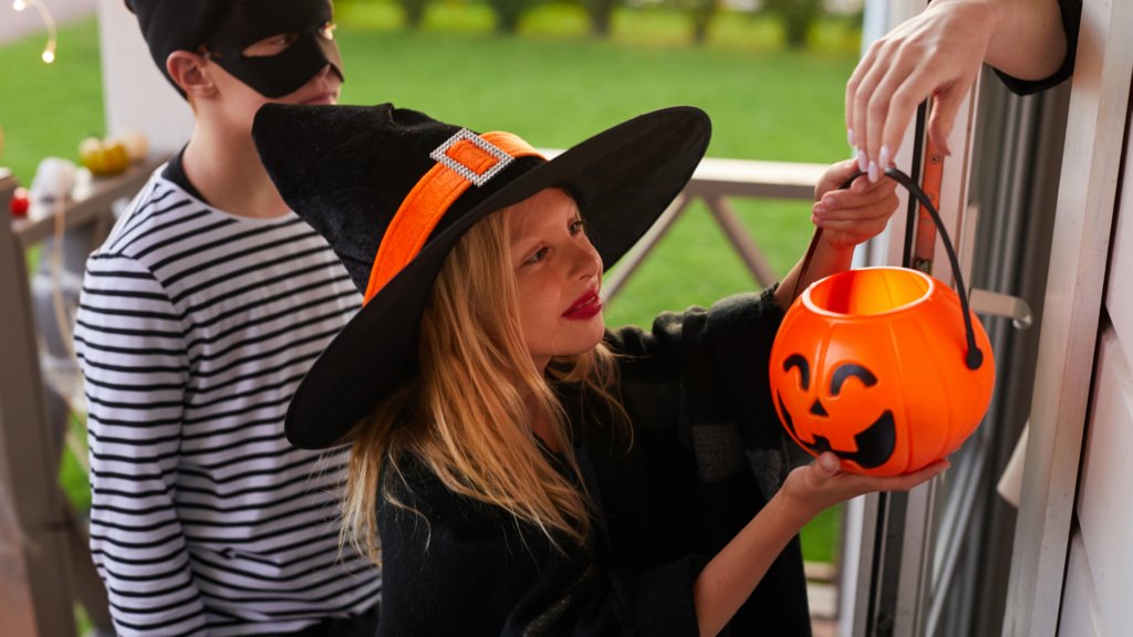 Two children in Halloween costumes hold candy buckets at a residential doorstep.