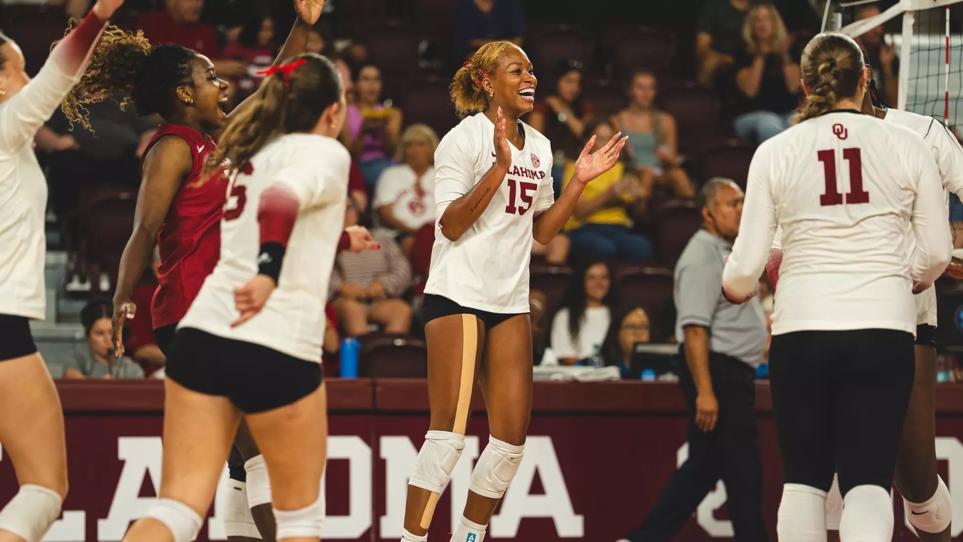 Female volleyball players in white and crimson uniforms celebrating on the court.