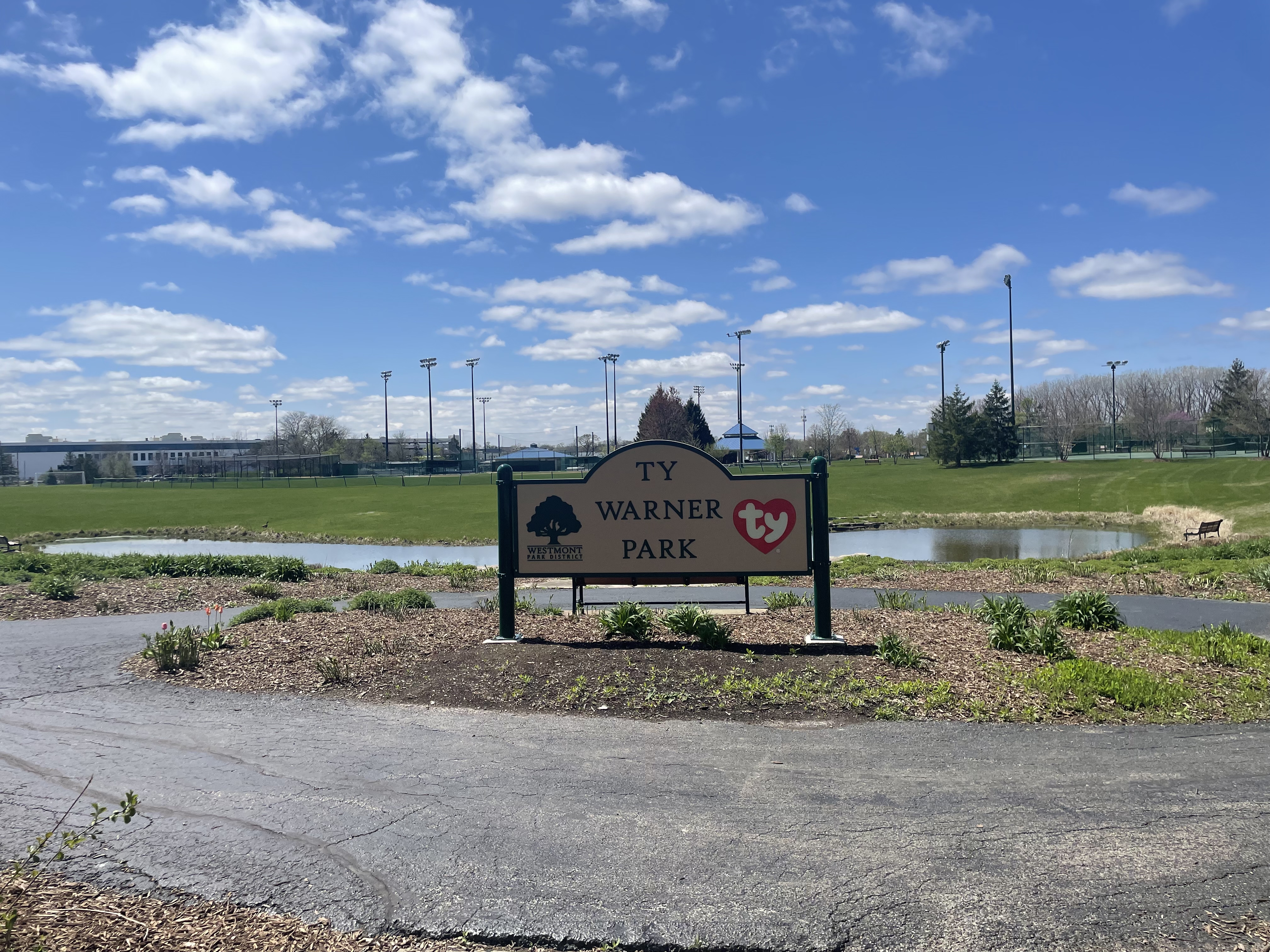 Park entrance sign beside a small pond with athletic field lights in the background.