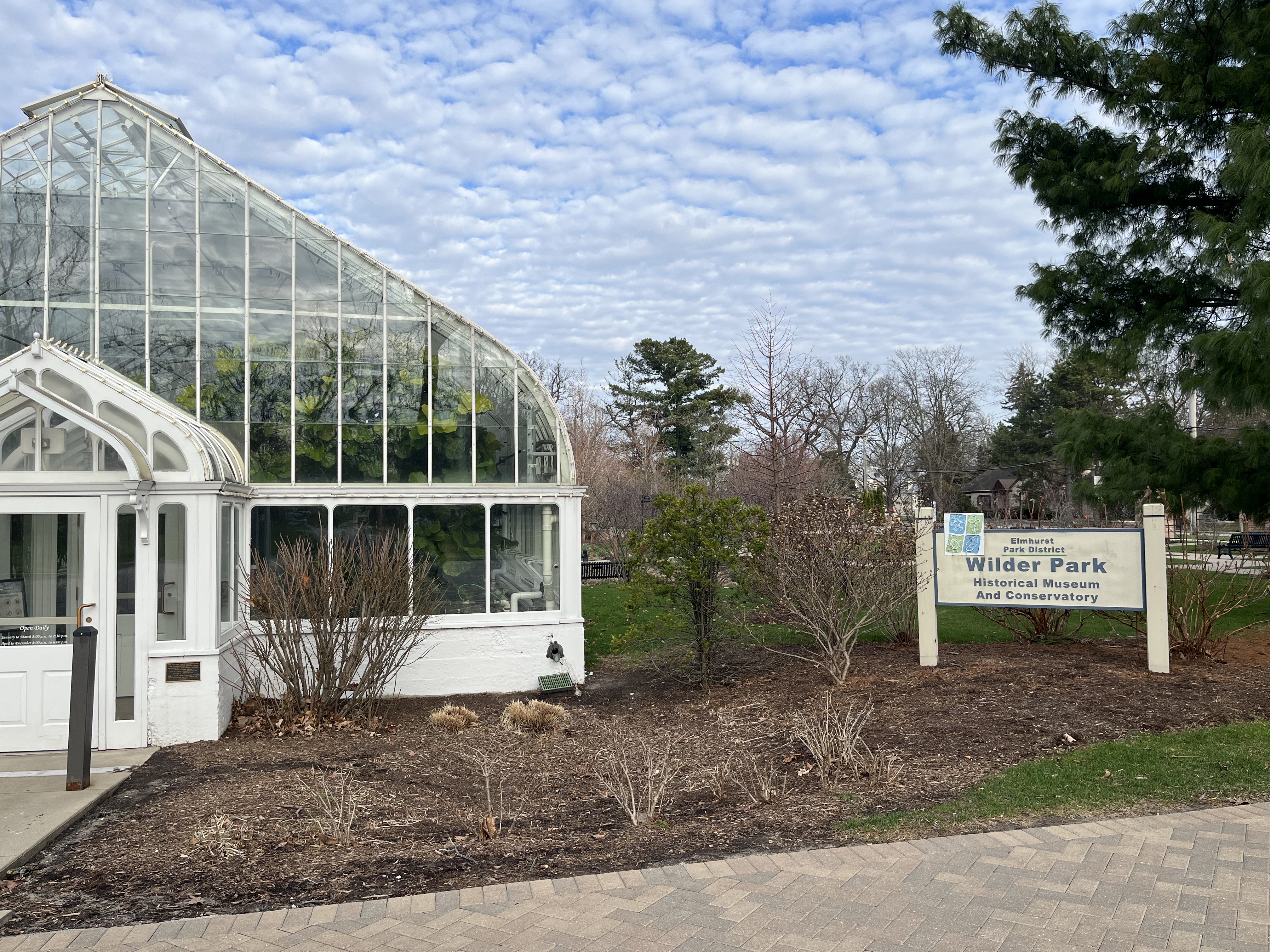 White Victorian-style glass conservatory building with a sign in front, surrounded by bare shrubs.